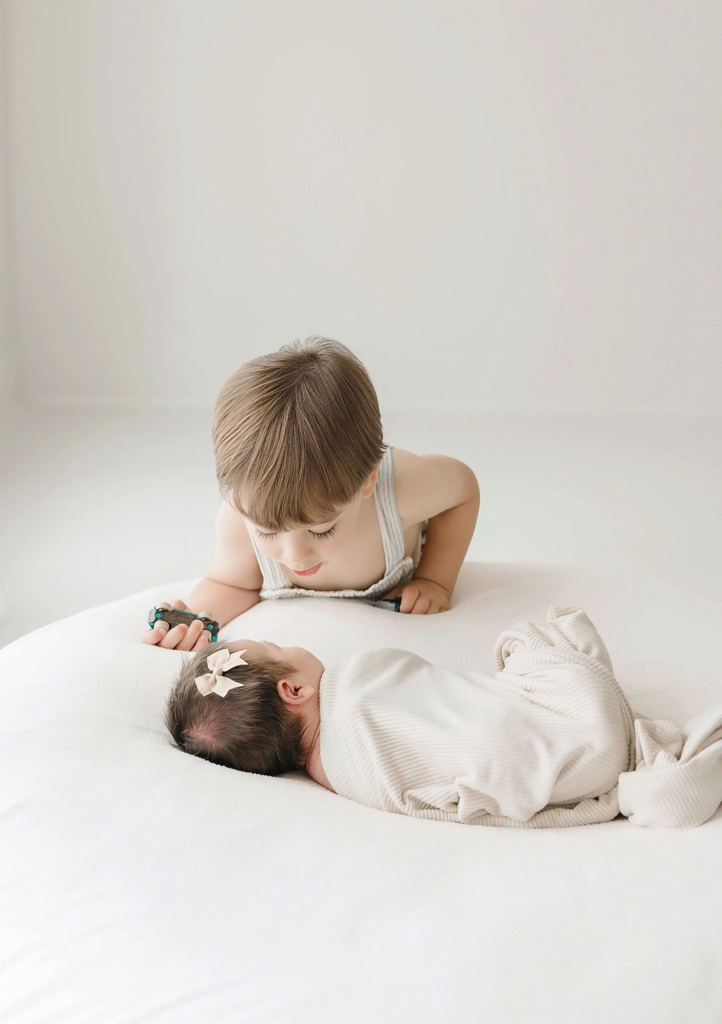 A young boy with brown hair, wearing a striped sleeveless shirt, leaning over a sleeping baby with dark hair, a cream bow, and striped clothing, on a white bed in a softly lit room. Baton Rouge Maternity Newborn and Family Photographer 