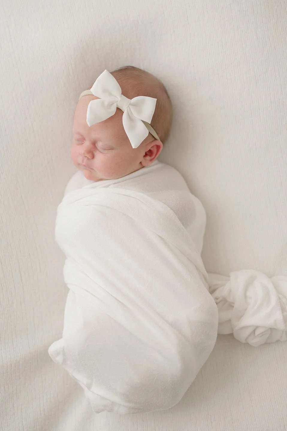 A sleeping newborn baby wrapped in a white blanket, wearing a white bow headband, resting against a light-colored background.
