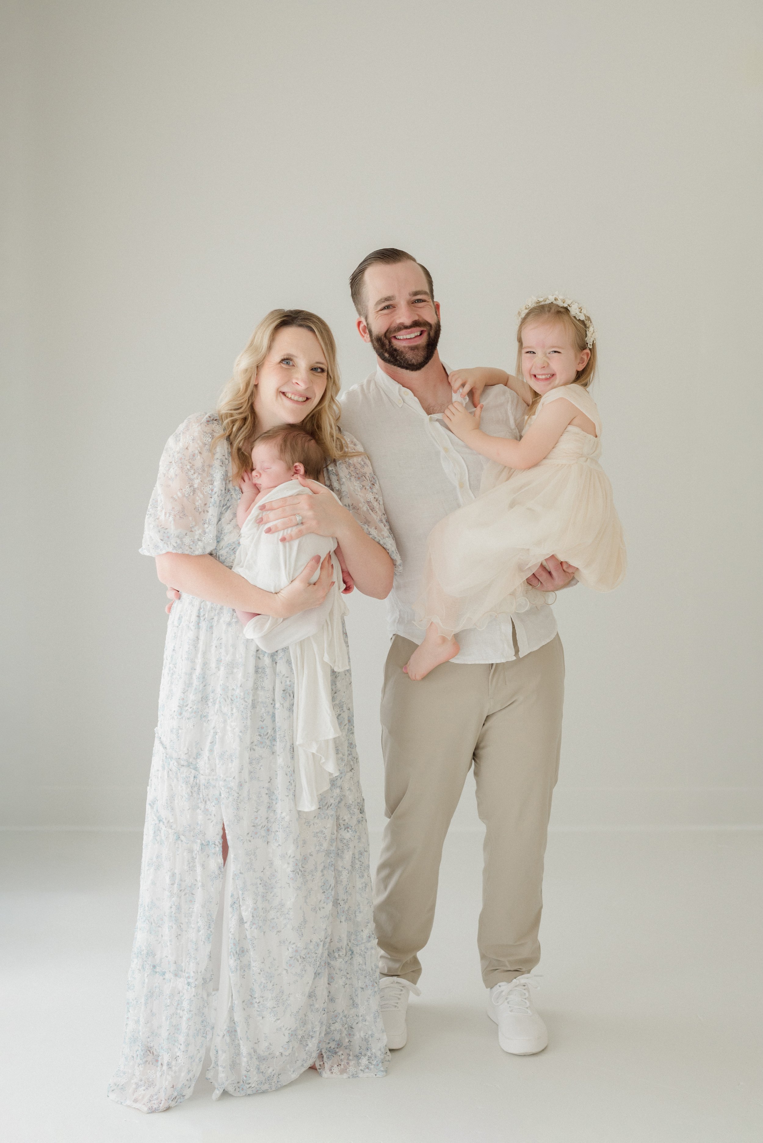 Family of four: mother, father, and two young daughters, smiling and posing together in a bright, neutral-colored studio. Baton Rouge Maternity Newborn and Family Photographer 