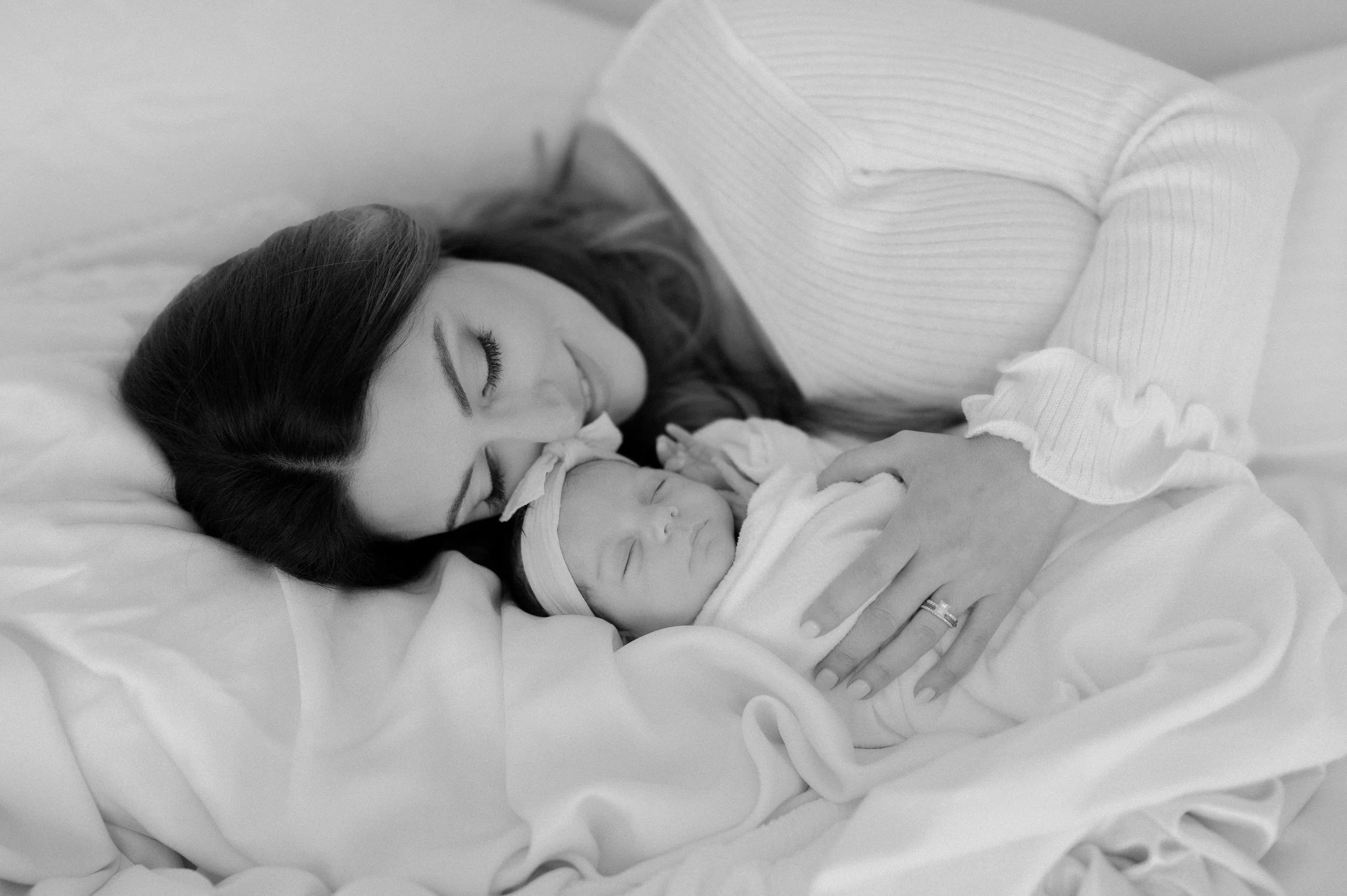 A woman with dark hair lying on a bed, tenderly holding and kissing a sleeping newborn baby wrapped in a blanket.