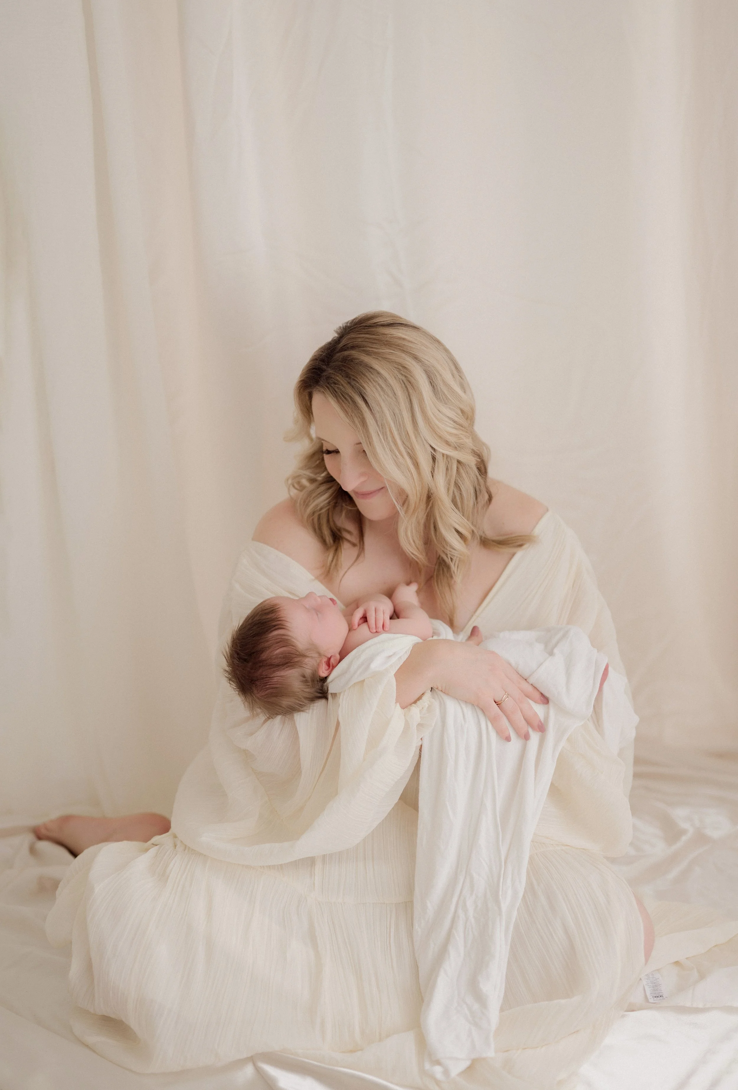 A woman with blonde wavy hair, wearing a cream-colored dress, sits on a bed holding a newborn baby wrapped in a white blanket, looking down at the baby with a gentle smile. Baton Rouge Maternity Newborn and Family Photographer 