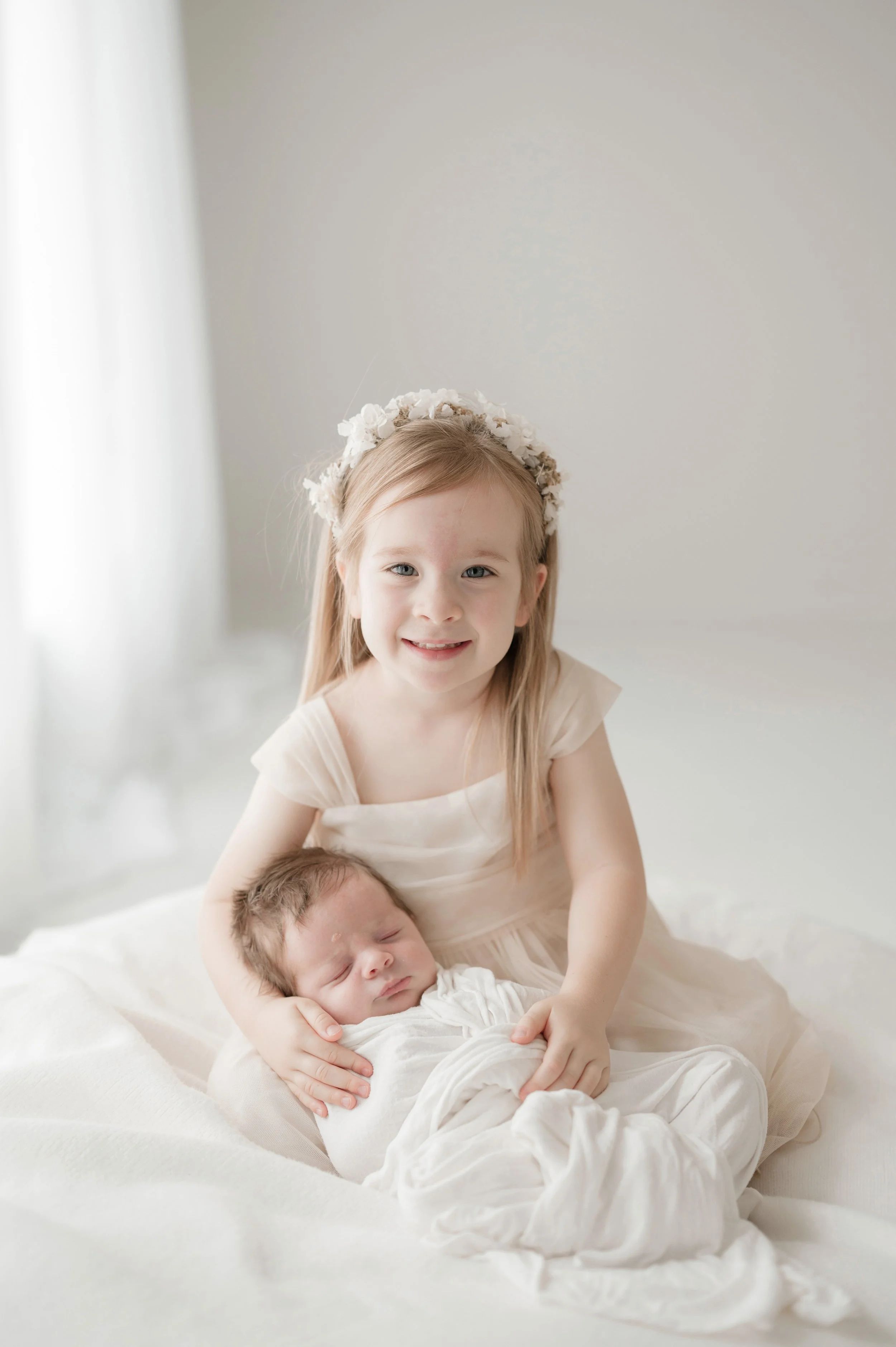 A young girl with a flower crown sitting on a bed and holding a sleeping newborn baby. Baton Rouge Newborn Photographer 