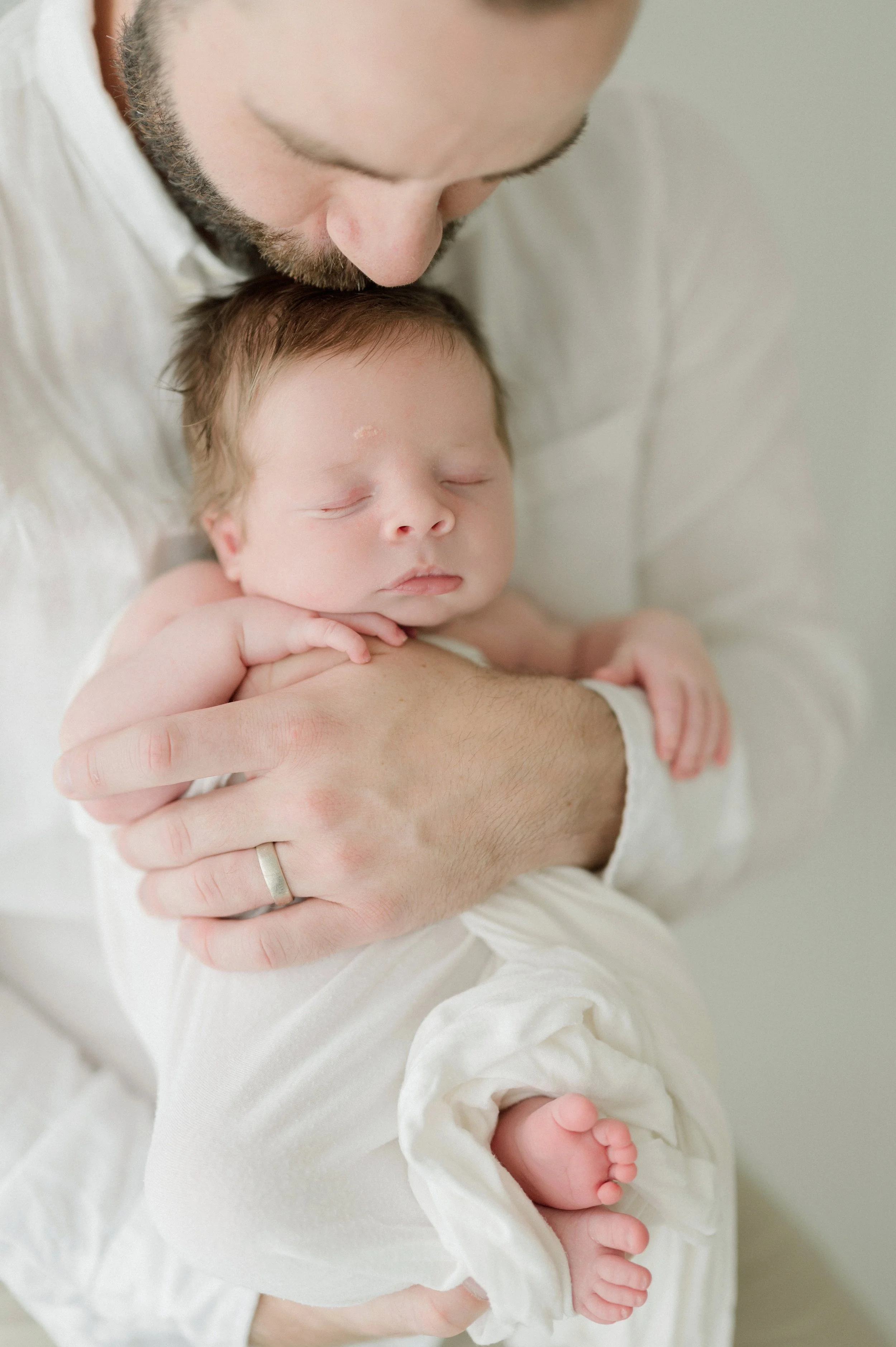 A man holding a sleeping newborn baby with a slight smile, wearing a white shirt, against a light background. Baton Rouge Maternity Newborn and Family Photographer 