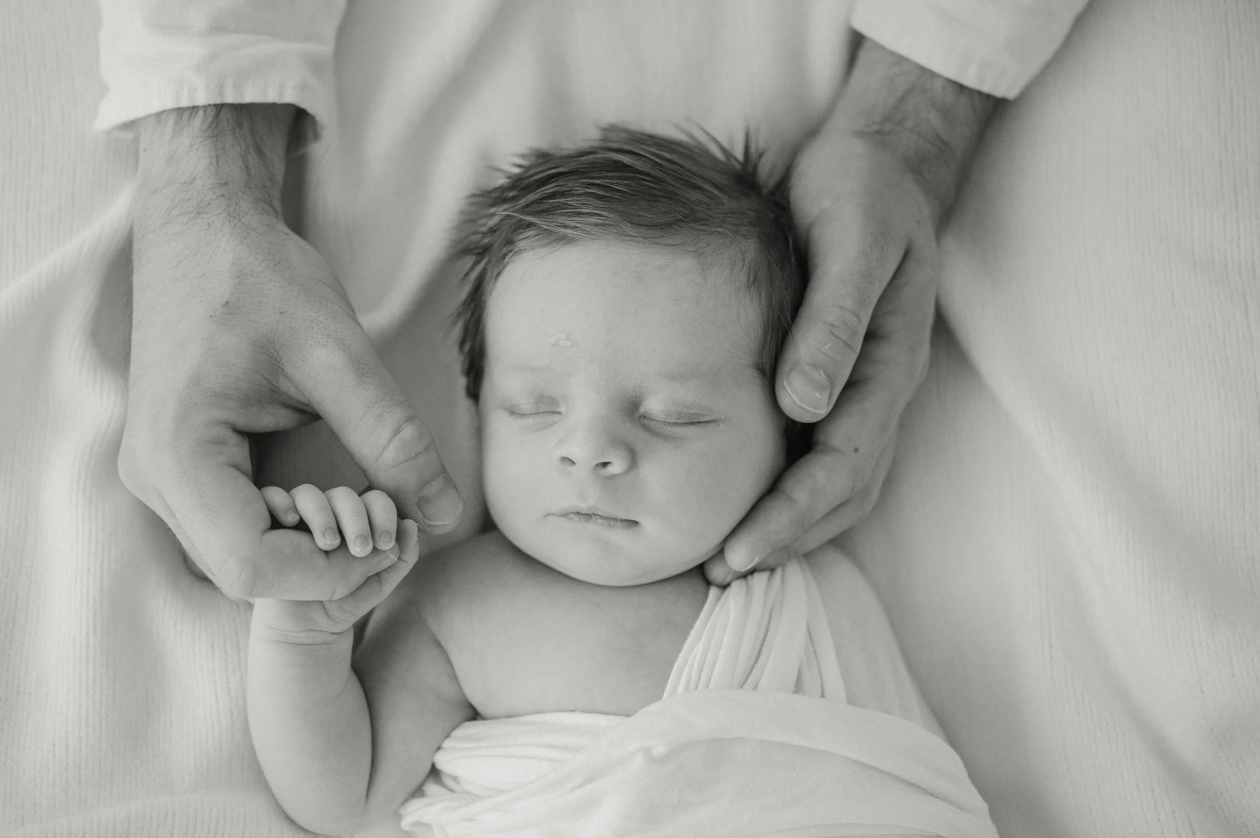 Black and white photo of a sleeping baby with closed eyes, surrounded by adult hands gently holding the baby's head and hand.