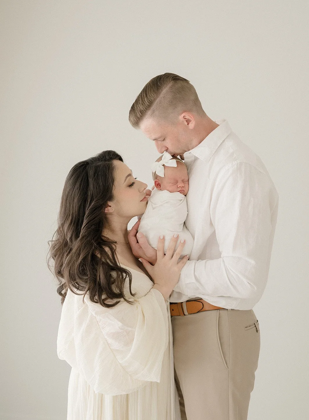 A woman and a man holding a newborn baby together in a neutral-colored background.