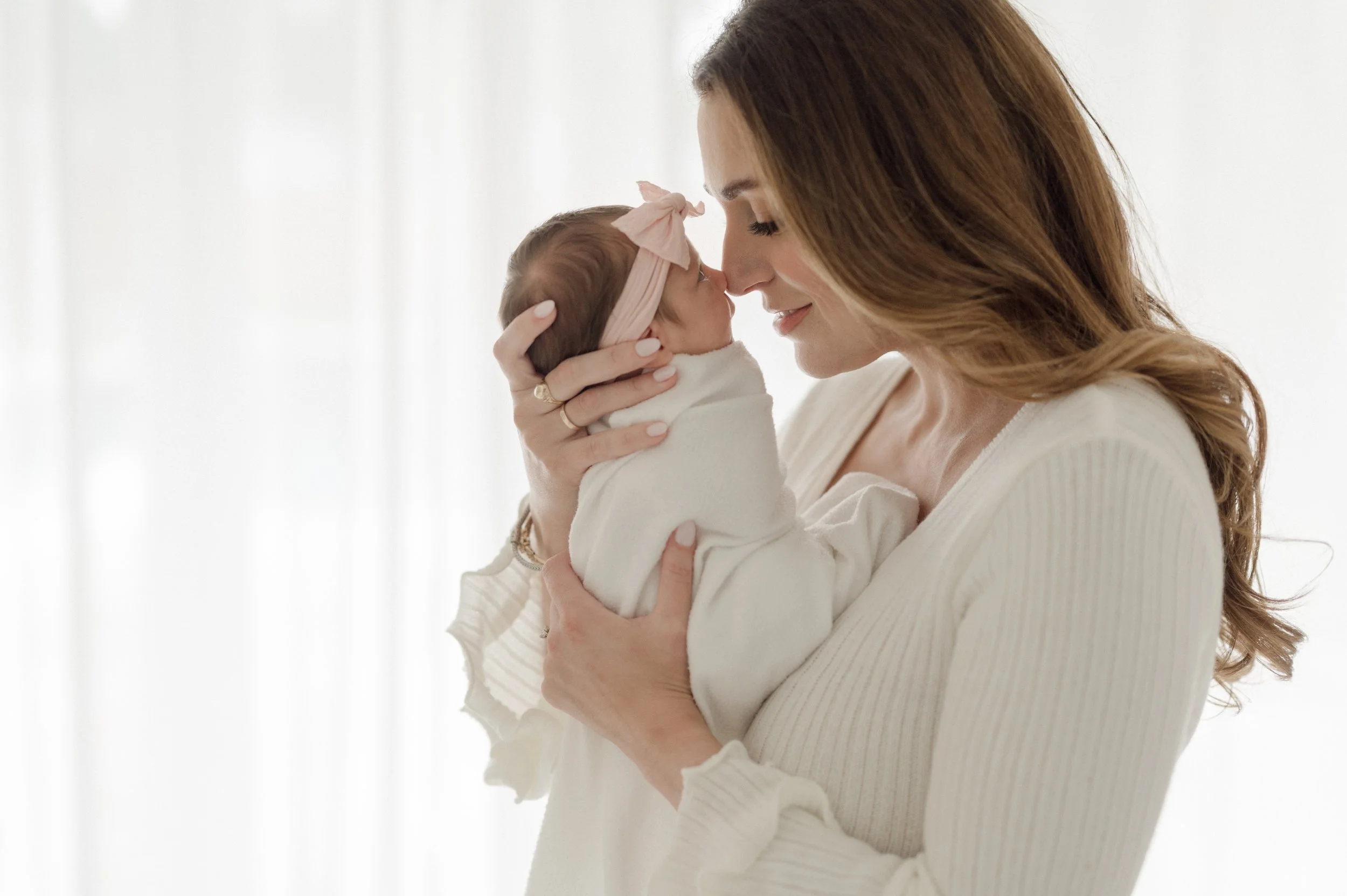 A woman with long brown hair holding a baby close, touching noses in a tender moment indoors with a white background.