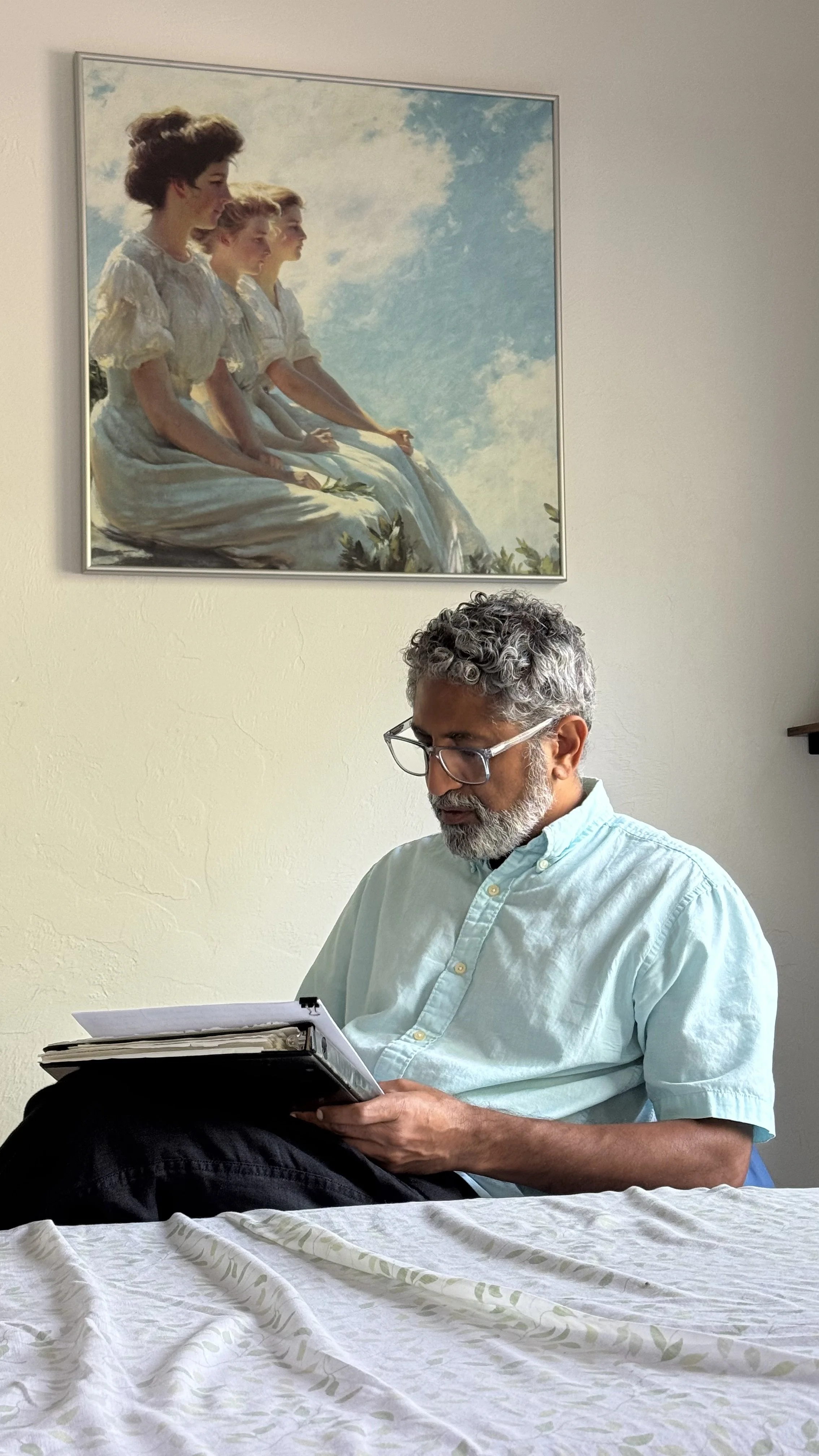 A man with gray curly hair, glasses, and a beard is sitting at a table, reading a folder. Behind him is a wall with a hanging painting of four women in white dresses sitting side by side, with a cloudy sky in the background.