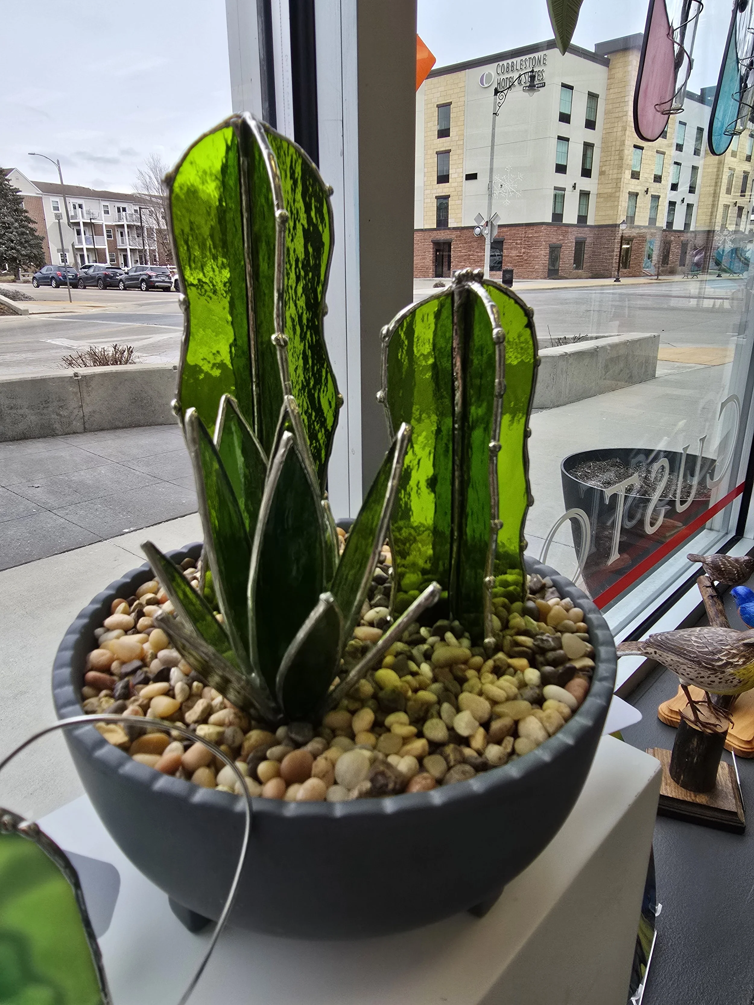 Decorative cactus made of green stained glass pieces in a black pot filled with small rocks, placed on a white surface near a window with views of buildings and street outside.