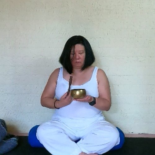 Debbie sitting in a cross-legged position on a bolster. She is holding a Tibetan singing bowl in her hands.