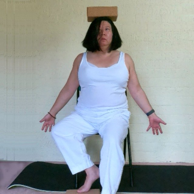 Debbie sitting in a chair doing the tree posture. She is resting one foot on a block and balancing another yoga block on her head.