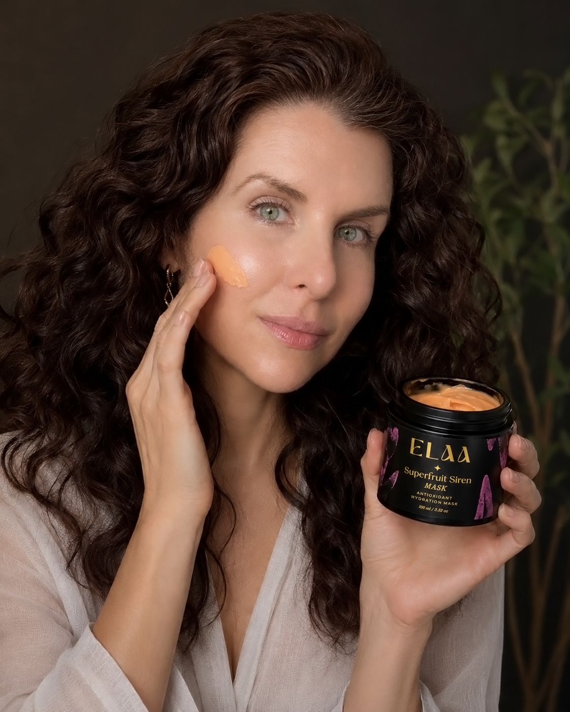 A black tray with skincare products, including ELAA Golden Sunrise Cleanser and Facial Oil, surrounded by flowers, leaves, and crystals, set against a dark background.