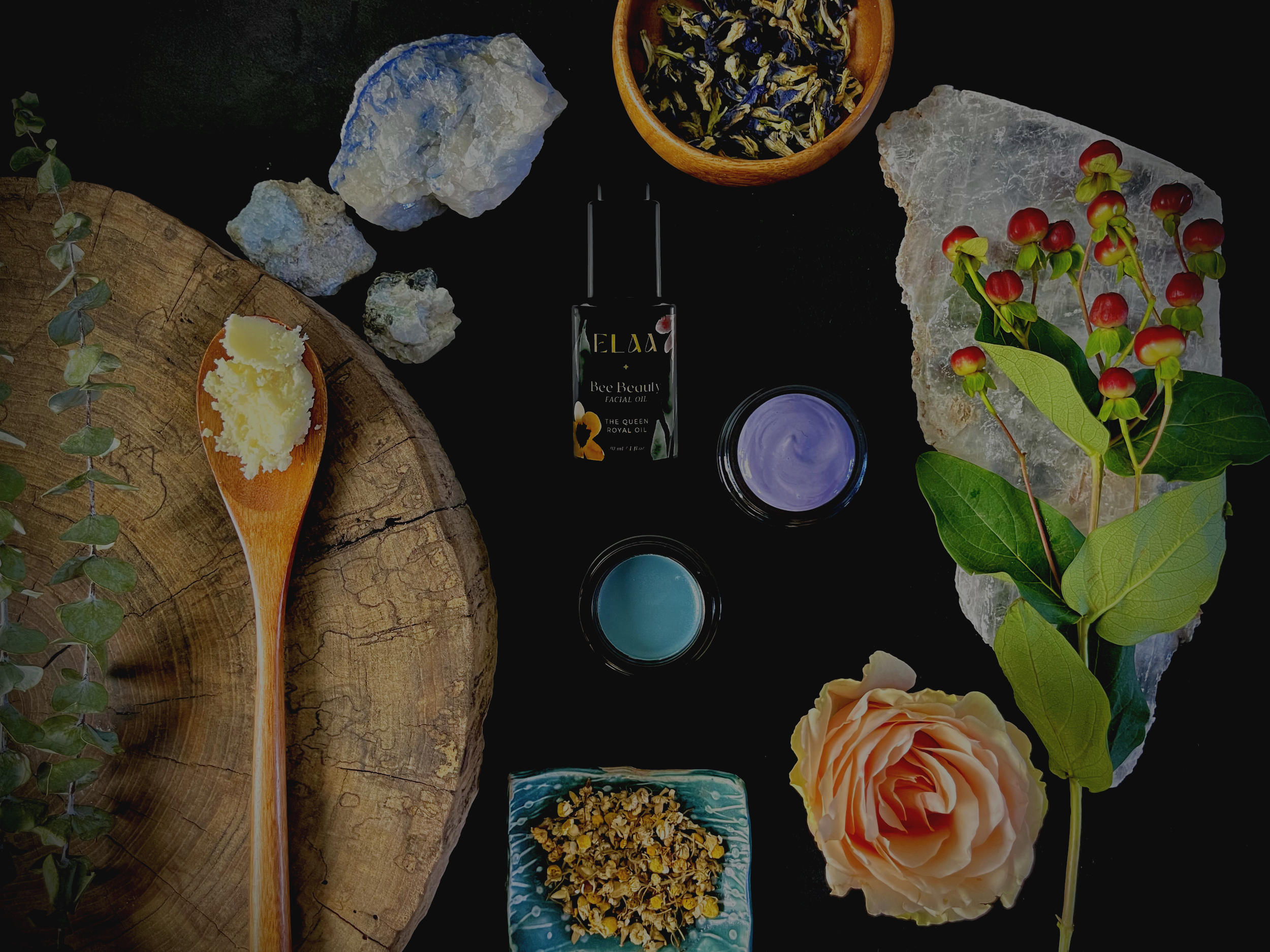 Skincare products, including a black bottle labeled 'ELAA Glow' and three small jars with pink, purple, and green creams, is arranged on a wooden surface with dried herbs, a white flower, purple and white quartz stones, and a few dried rose hips.
