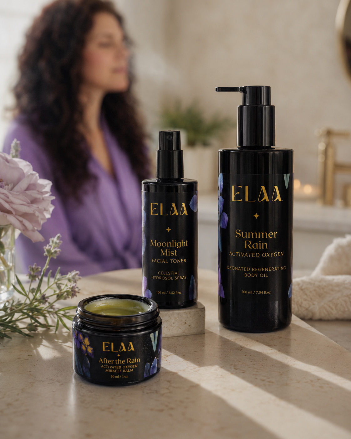 A bottle of ELAA Skincare Glazed Elixir Radiance Oil standing next to its packaging box, surrounded by flowers and petals with a textured green wall background.