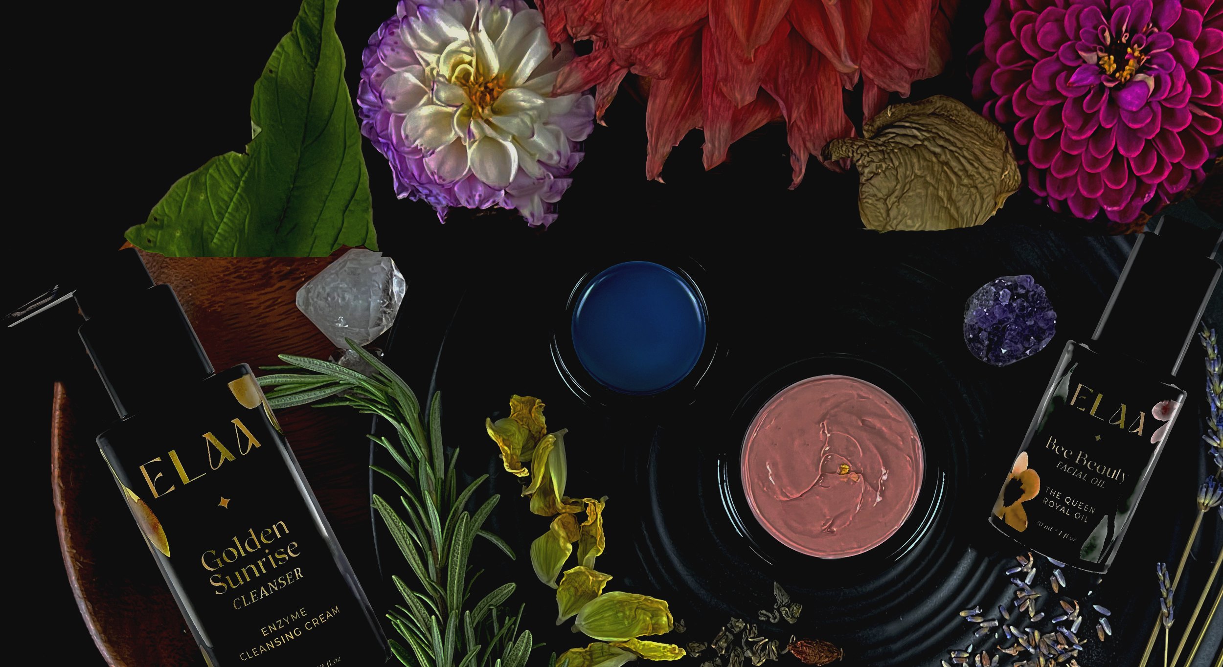 A black tray with skincare products, including ELAA Golden Sunrise Cleanser and Facial Oil, surrounded by flowers, leaves, and crystals, set against a dark background.