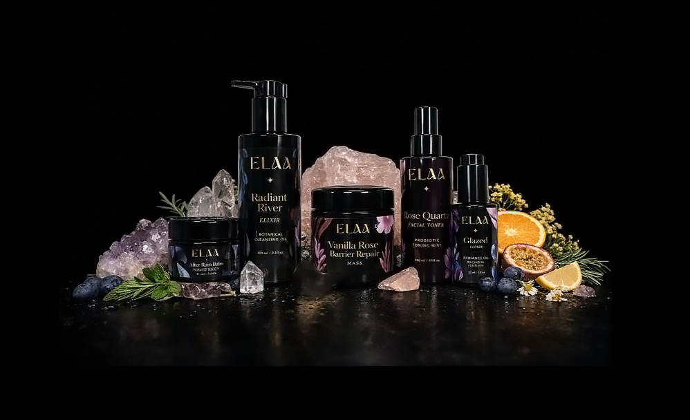 Collection of skincare products and aromatherapy elements on a black background, including bottles labeled ELAA Radiant River, Rose Quartz Facial Toning Mist, Glazed Radiance Oil, Vanilla Rose Barrier Repair Mask, and a small jar of ELAA After Rain Balm, surrounded by crystals, herbs, and citrus slices.