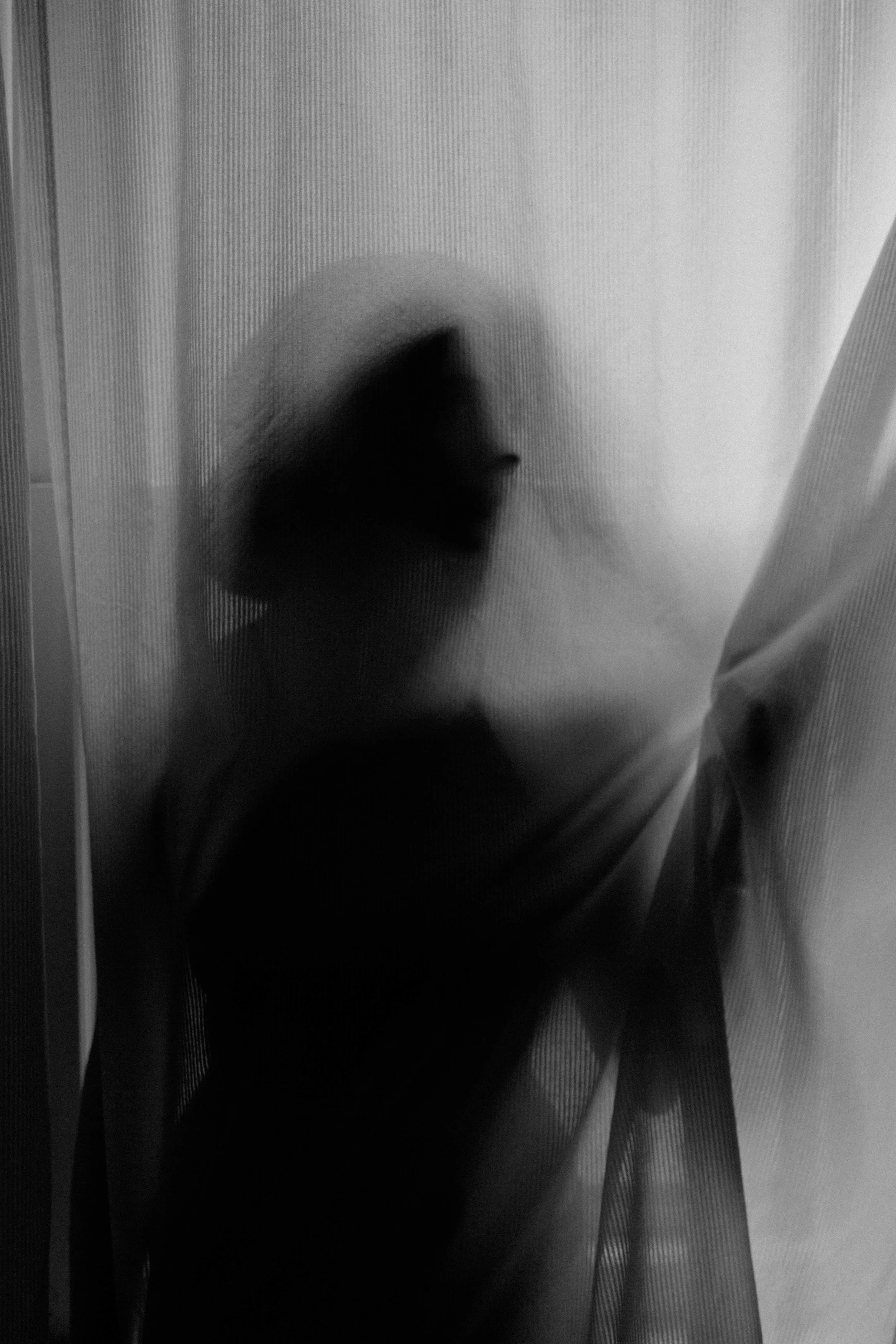 a black-and-white portrait of emma grimsley in a victorian airbnb in dayton, ohio. photograph by sarah rose walk of sarah rose photography. emma is standing behind a slightly sheer curtain, and she is looking to the side. emma is wearing a black lacy crop bralette.