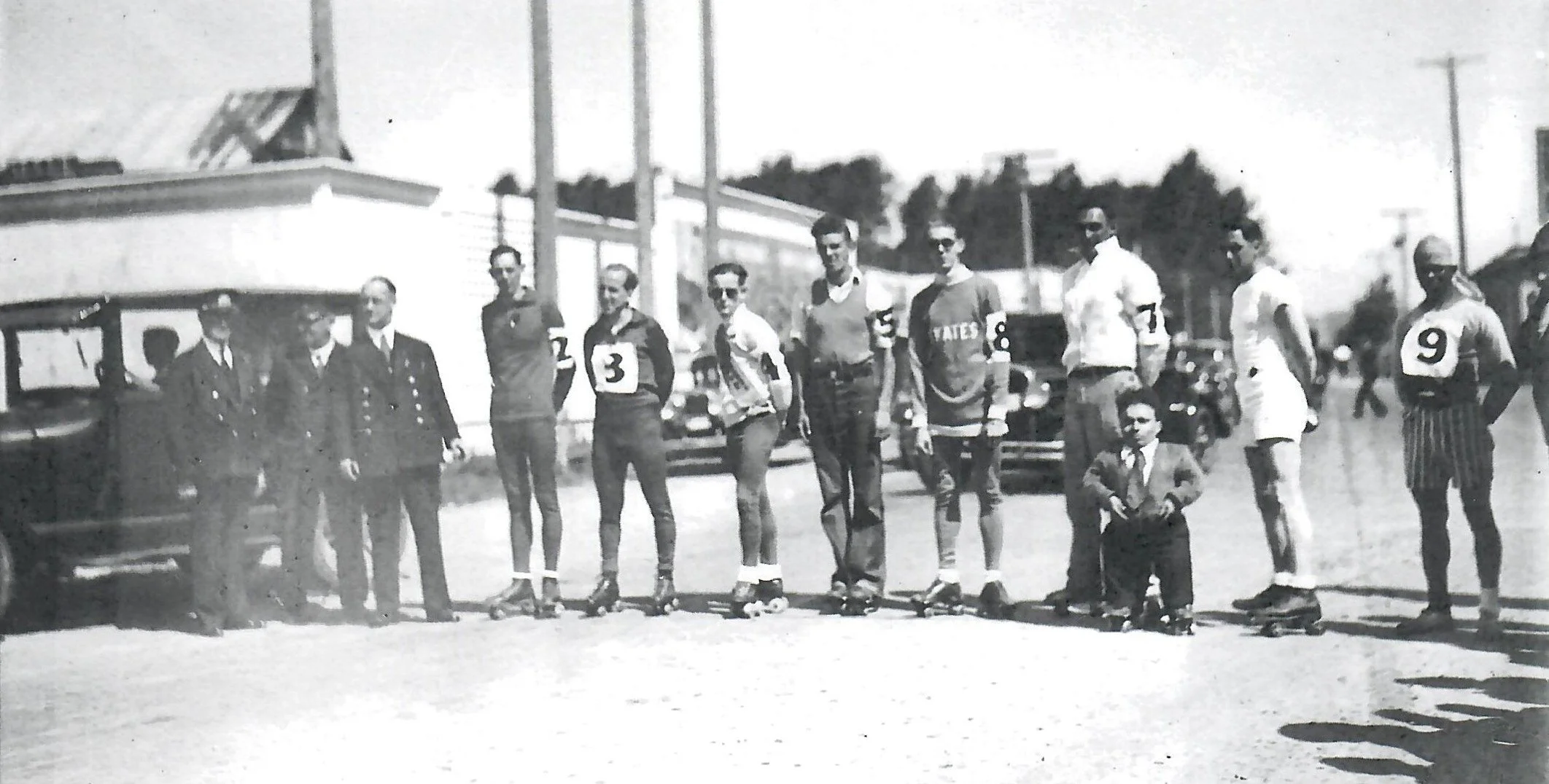 Black and white vintage photo showing line of men on rollerskates standing on road, they have numbers on as contestants in a race. They include a little person and a man of color.  Several policeman stand off to one side.
