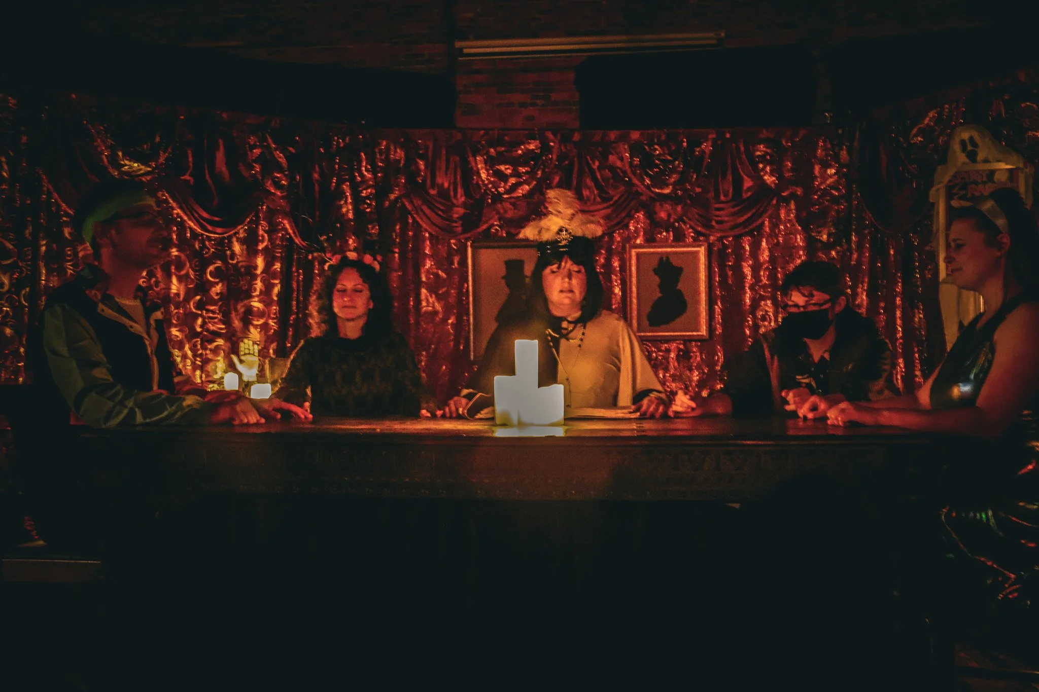 Five persons sit around a table holding hands in a dimly lit room with candles and gaudy curtains behind them , holding a seance