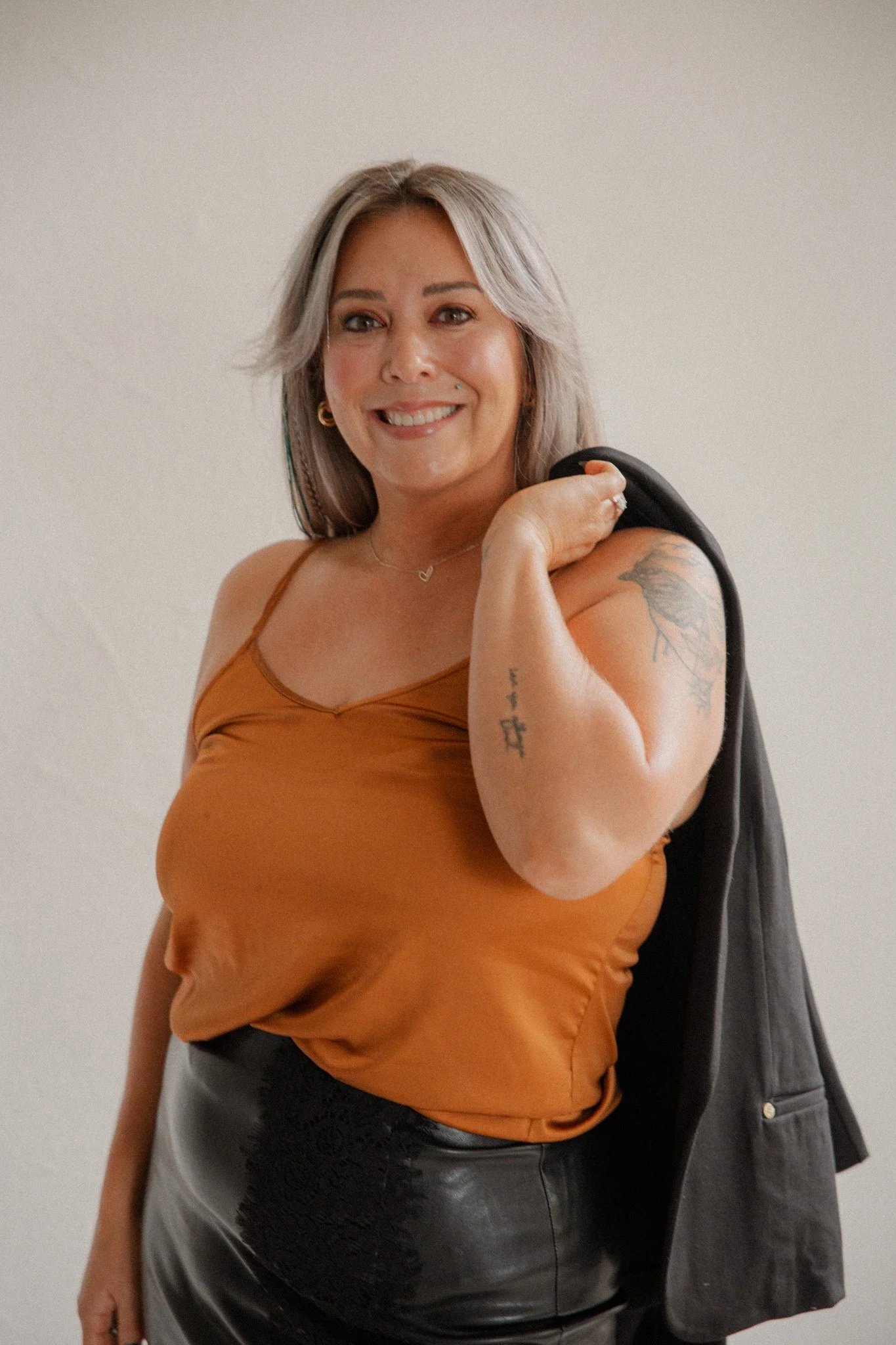 Kaitlyn Lakey, hair stylist at Modèle Salon in Oregon City, with silver-blonde hair, smiling in an orange tank top with a black blazer over her shoulder