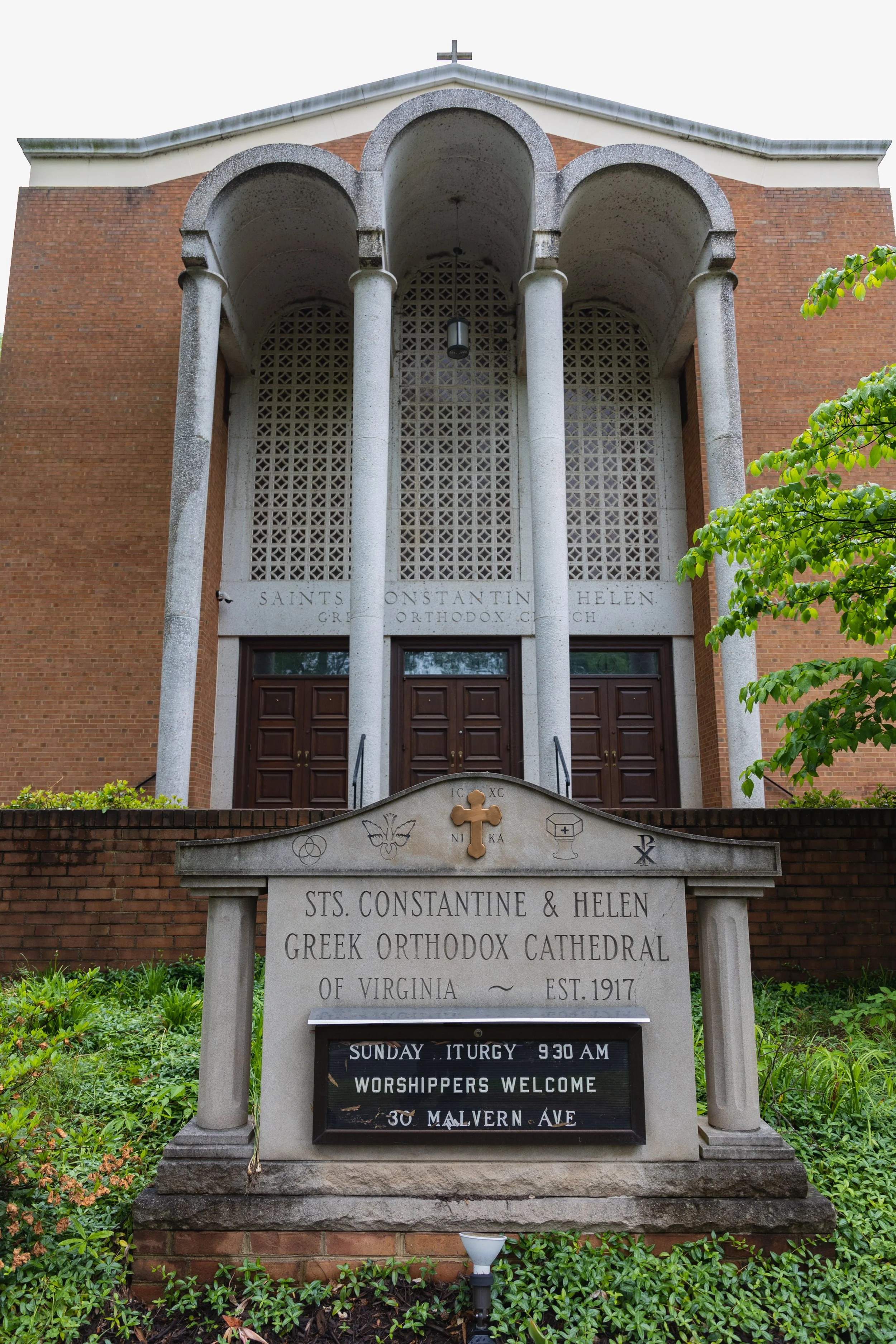 The Greek Orthodox cathedral named Sts. Constantine and Helen in Virginia, established in 1917, with brick walls, a decorative stone archway entrance, and a stone sign with religious symbols and service hours in front surrounded by greenery.