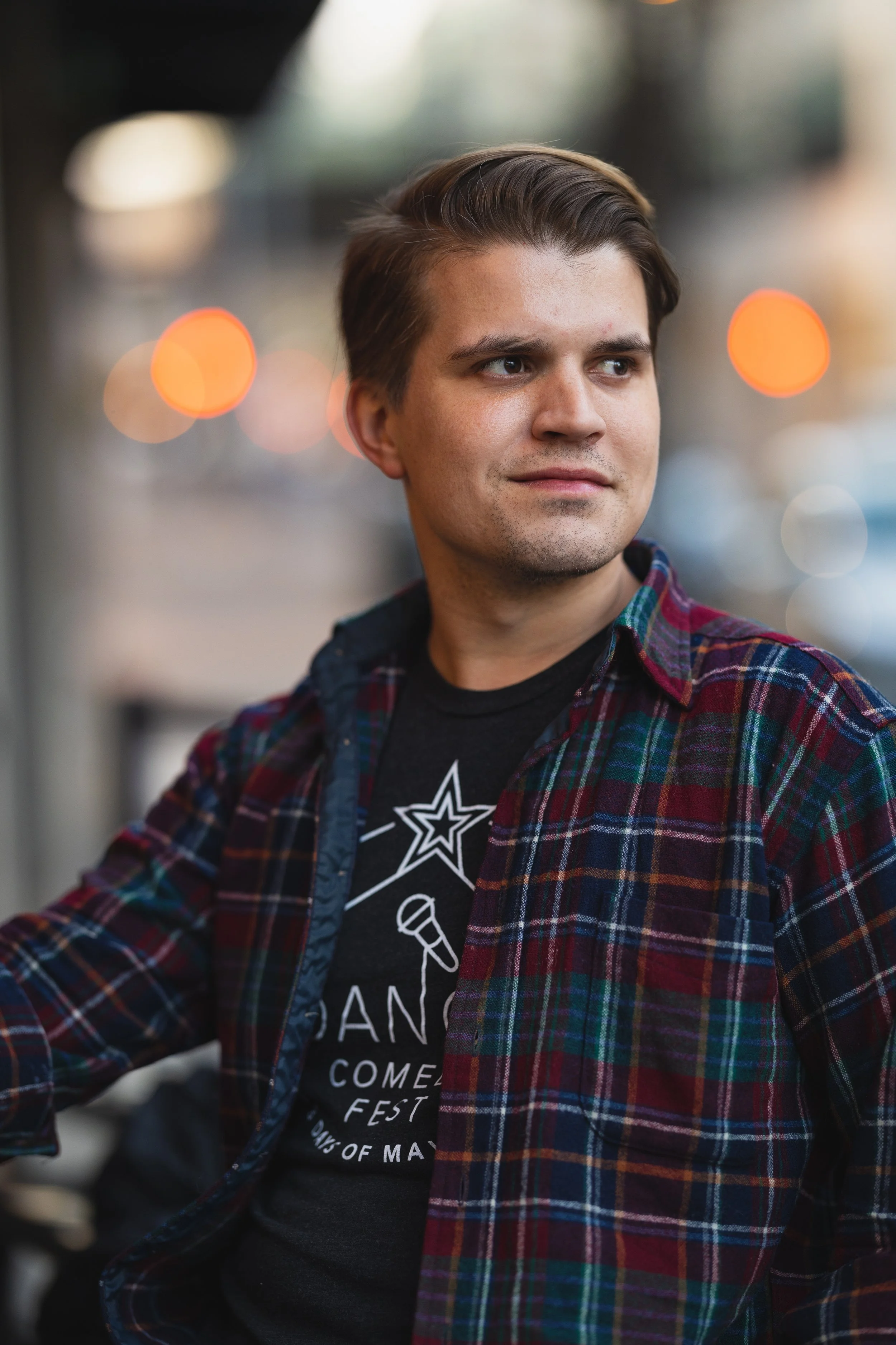 A young man with brown hair and light skin wearing a black comic-con T-shirt and a red, green, and blue plaid shirt, looking to his right with a slight smile. The background is blurred with bokeh lights.