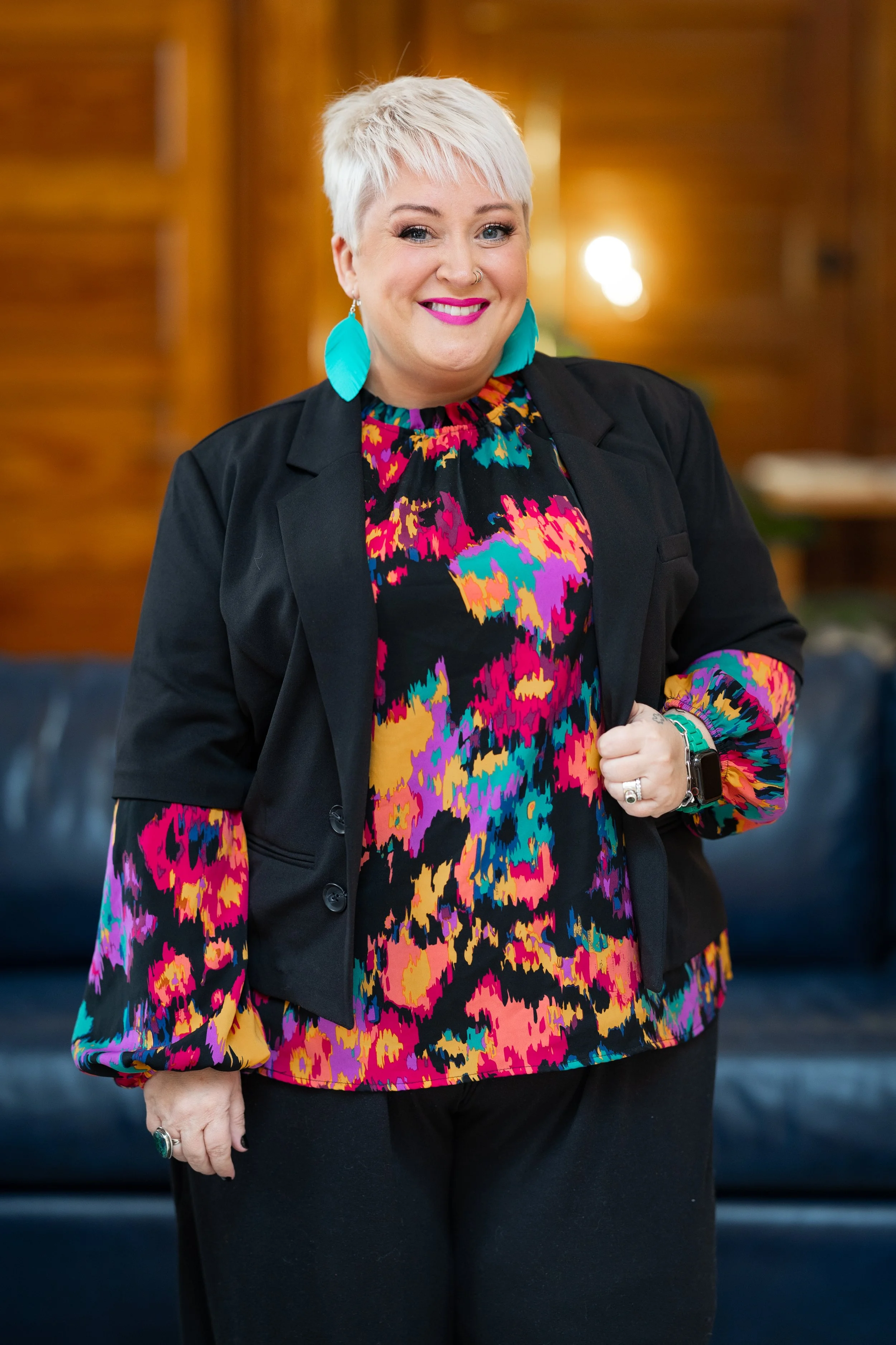A woman with short white hair, wearing a colorful patterned dress, black blazer, teal earrings, pink lipstick, and a nose ring, smiling inside a warmly lit room.