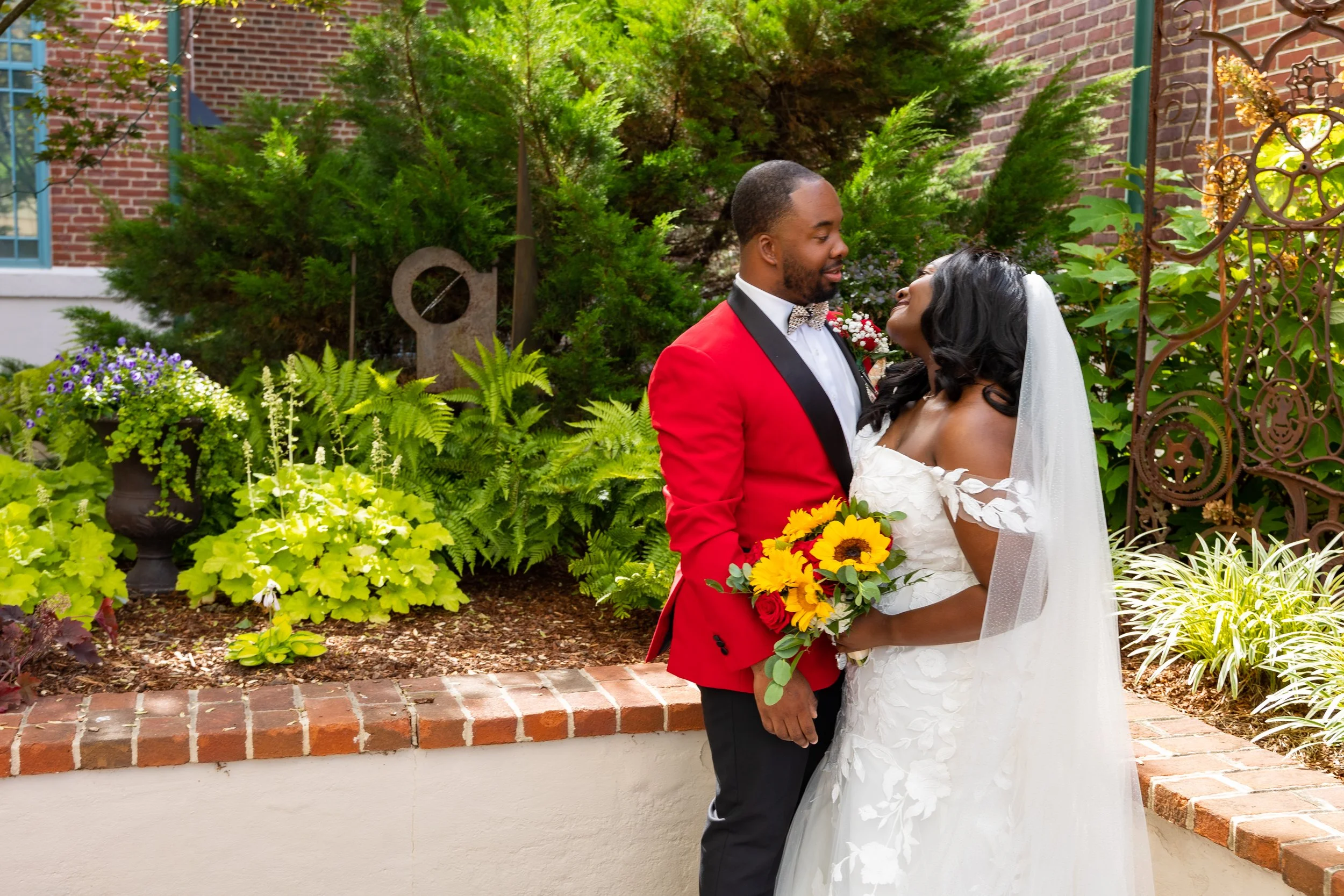 A couple dressed in wedding attire, standing outdoors near a brick building, surrounded by lush green plants and flowers, sharing a tender moment.
