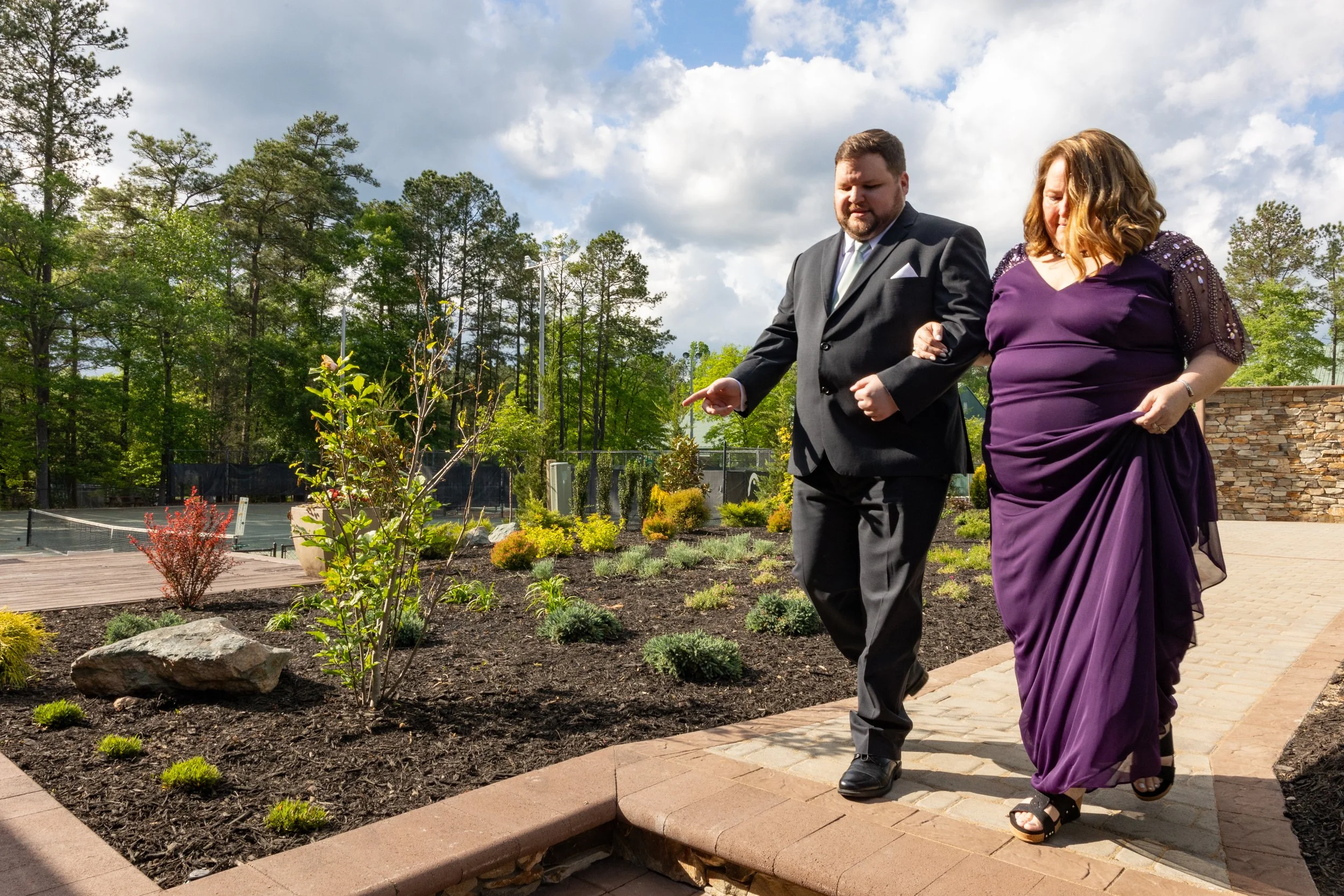 A man in a black suit and a woman in a purple dress walking arm in arm along a garden pathway on a partly cloudy day.