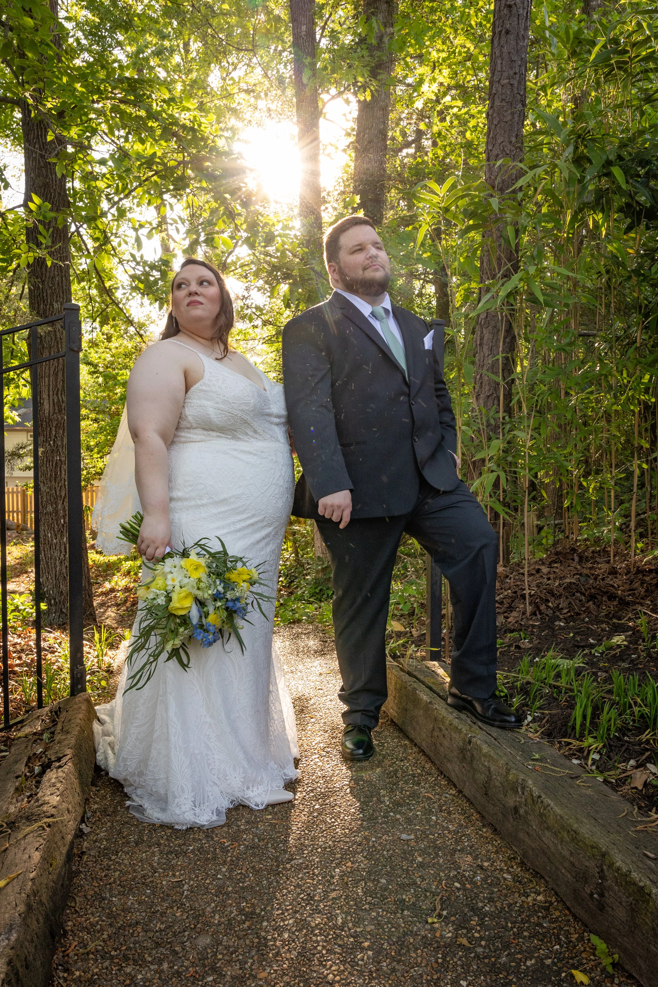 A bride and groom standing outdoors in a wooded area during a wedding ceremony, with sunlight filtering through trees in the background. The bride is holding a bouquet of yellow and blue flowers, and the groom is wearing a black suit with a light gre