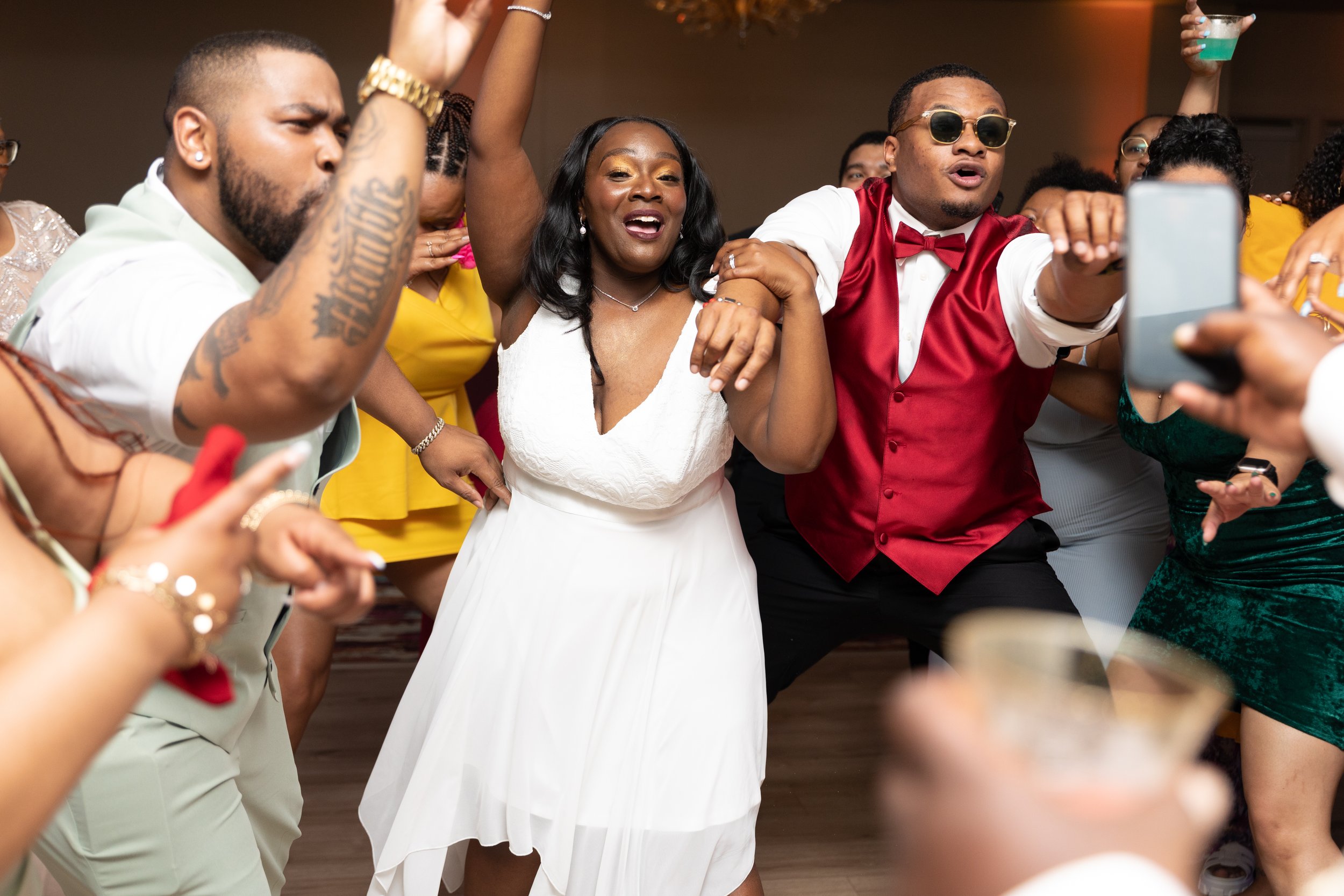 People dancing and celebrating at an indoor party, with many raising their hands and holding drinks, including a woman in a white dress and a man in sunglasses and a red vest.