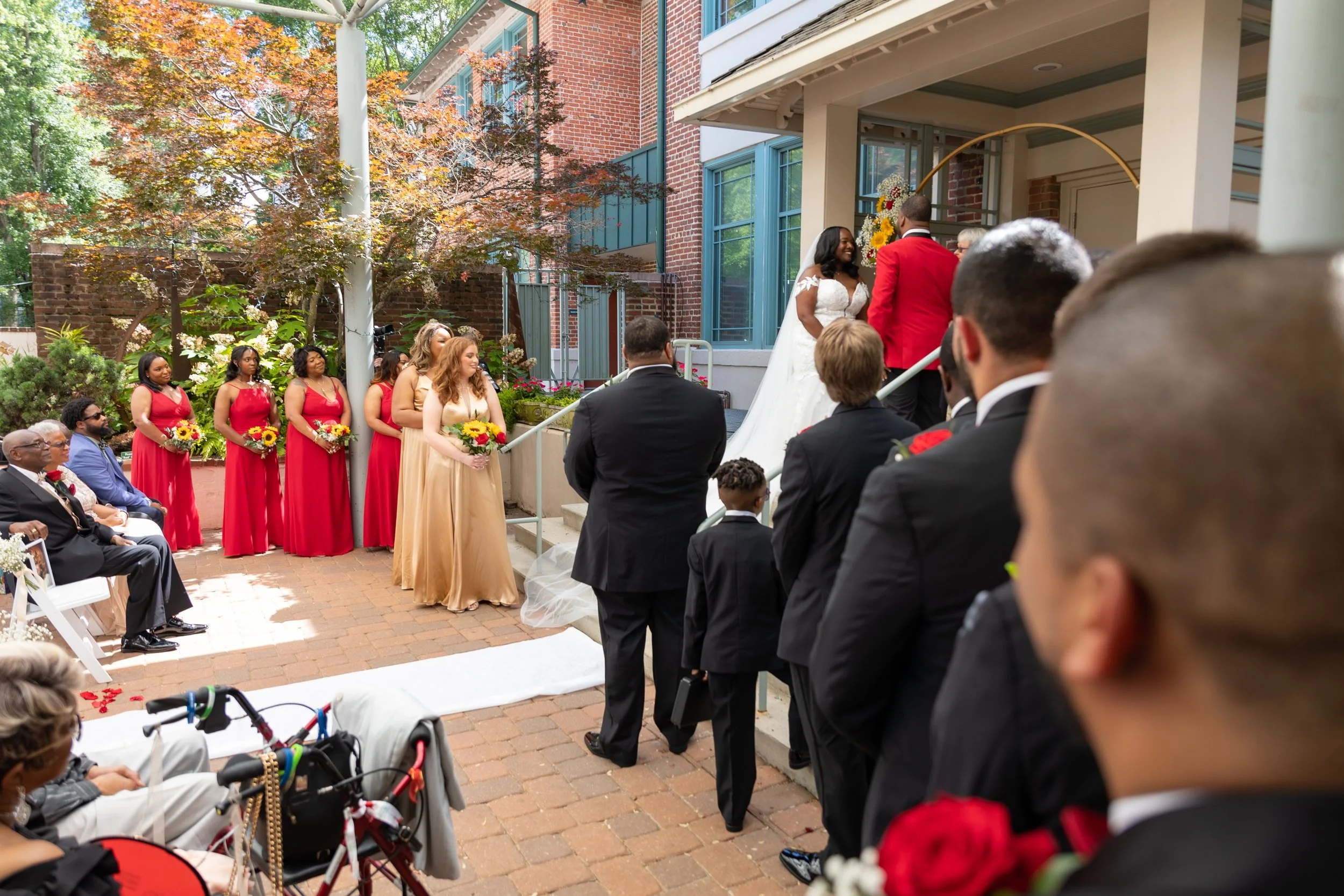 A wedding ceremony with the bride and groom standing outside a brick building on a porch, surrounded by guests. The bride is wearing a white wedding dress and the groom is in a red jacket. Bridesmaids in red dresses and guests are seated and standing