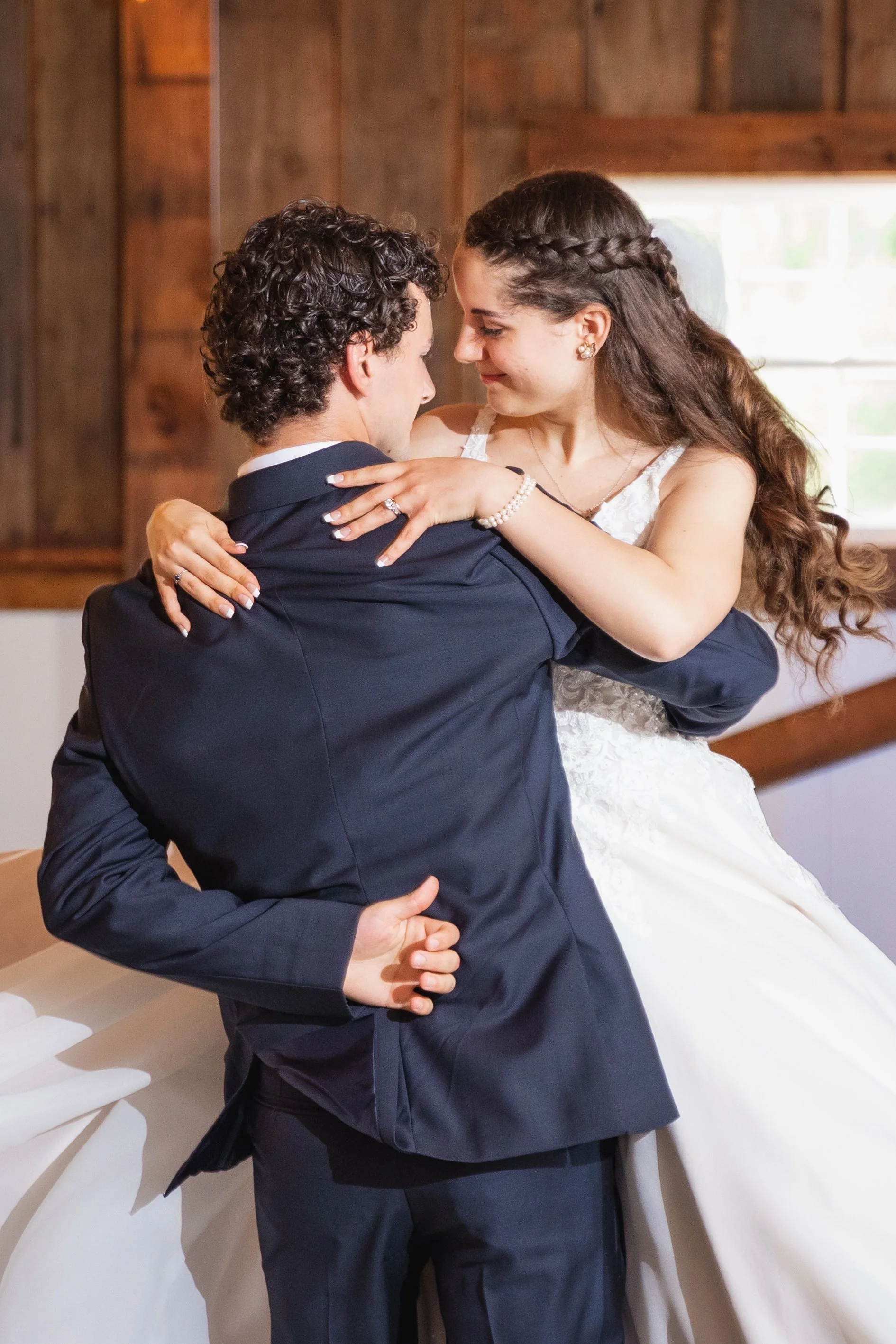 A couple dressed in wedding attire dancing closely inside a room with wooden walls and a window letting in natural light.