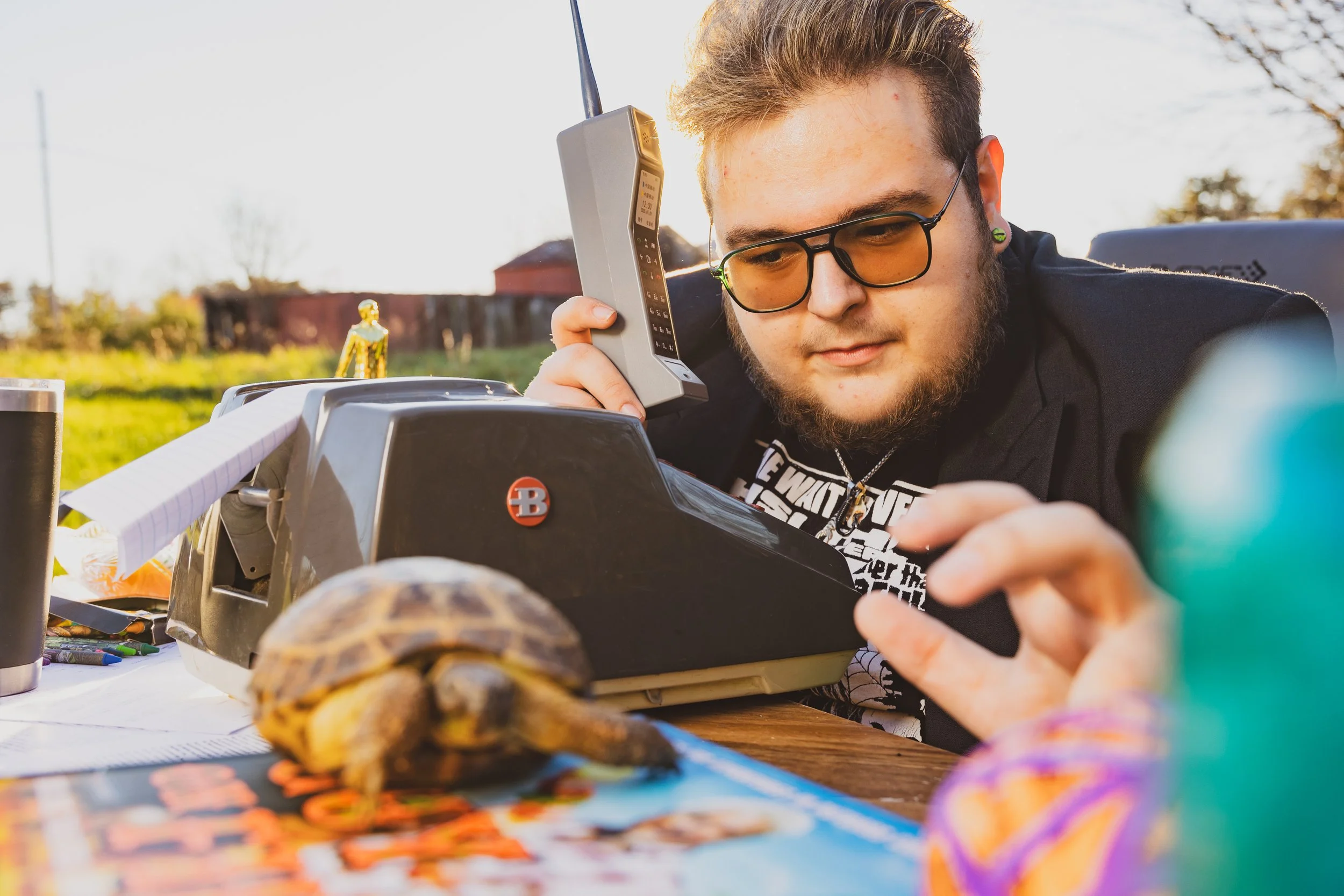 A young man with glasses and a beard is speaking into a phone while sitting outdoors at a table with tortoises, papers, and pens, during what appears to be a sunny day.