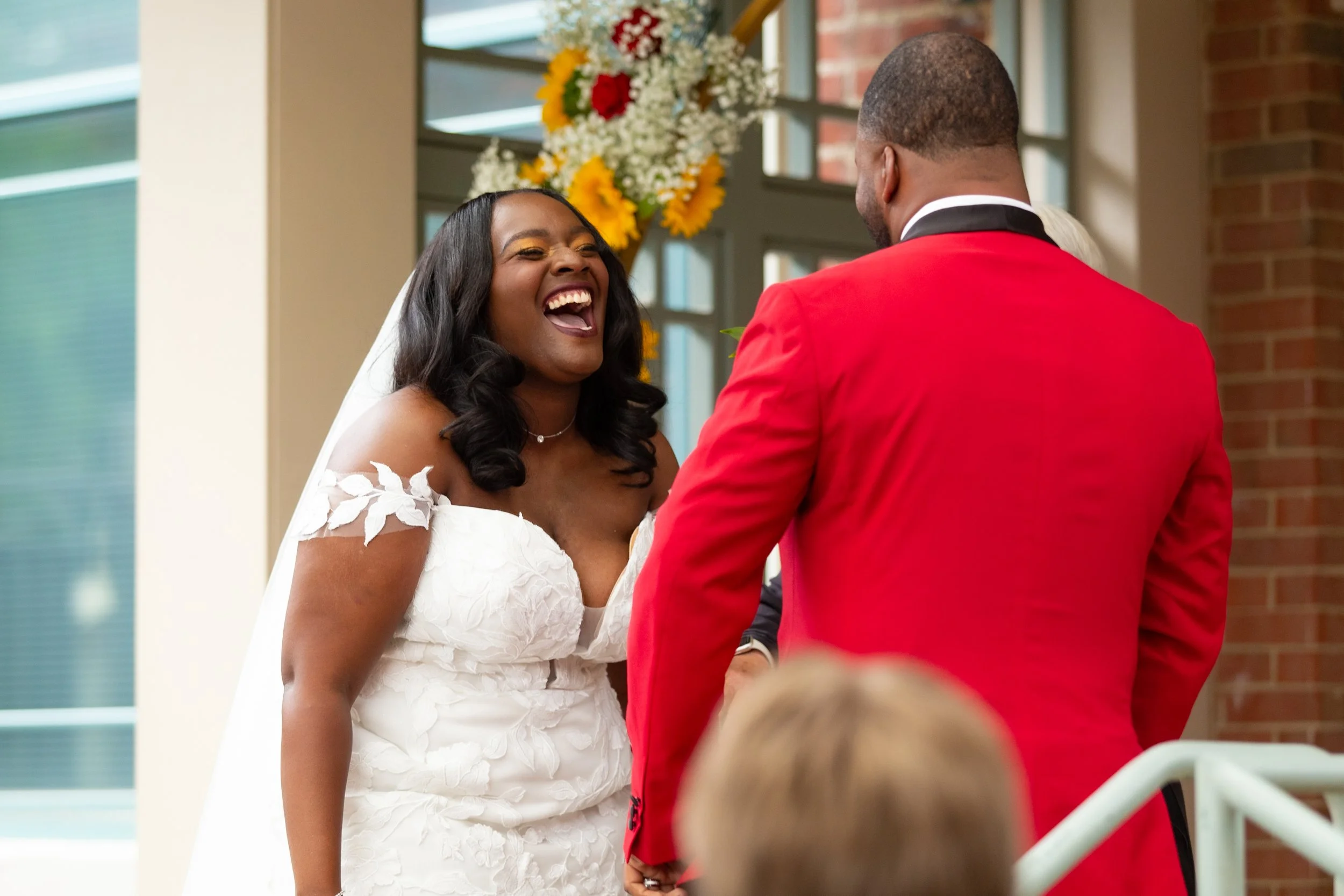 A bride with long dark hair and a white wedding dress laughing and holding hands with a man in a red suit.