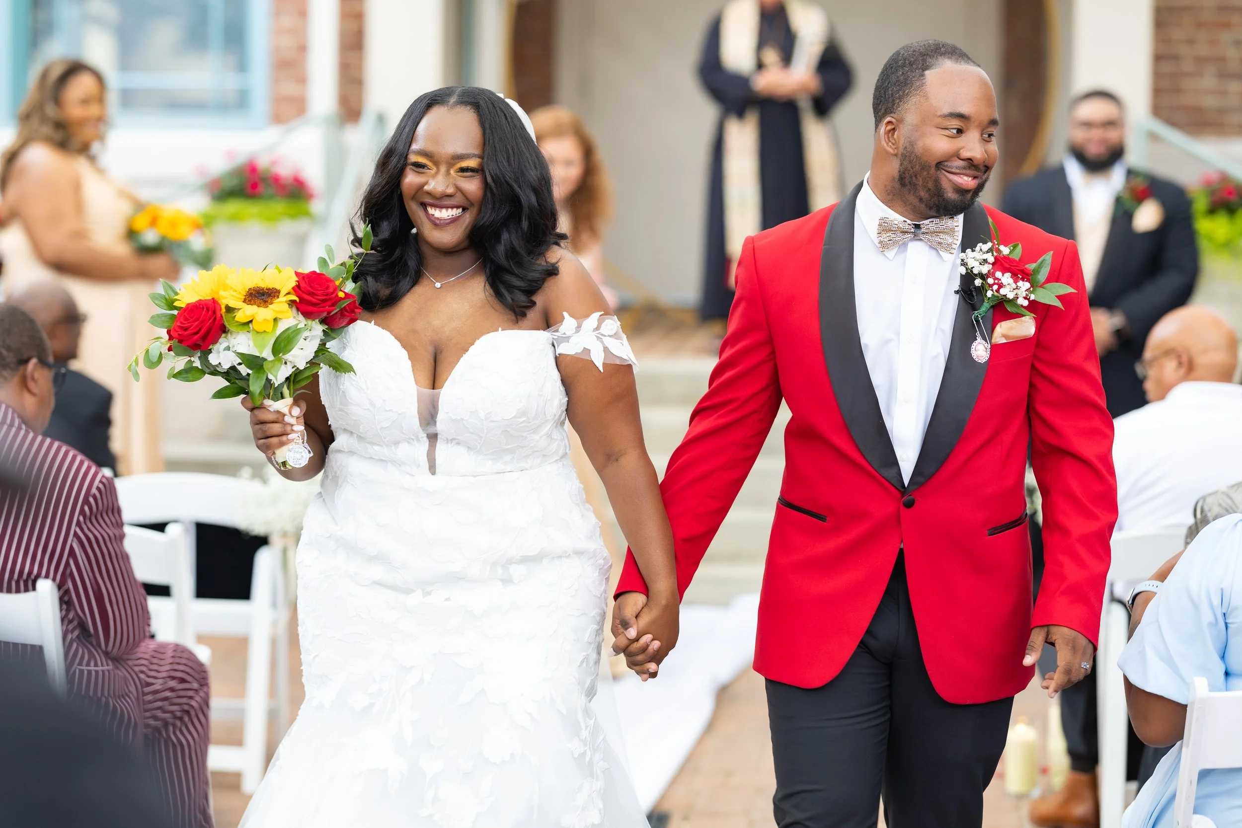A bride and groom holding hands during their wedding ceremony, walking down the aisle with guests seated on either side. The bride is smiling and holding a bouquet of red, yellow, and white flowers; she is wearing a white wedding gown. The groom is wearing a red tuxedo jacket with a boutonniere, a bow tie, and black pants. The background shows a decorated venue with some guests and a officiant standing behind.