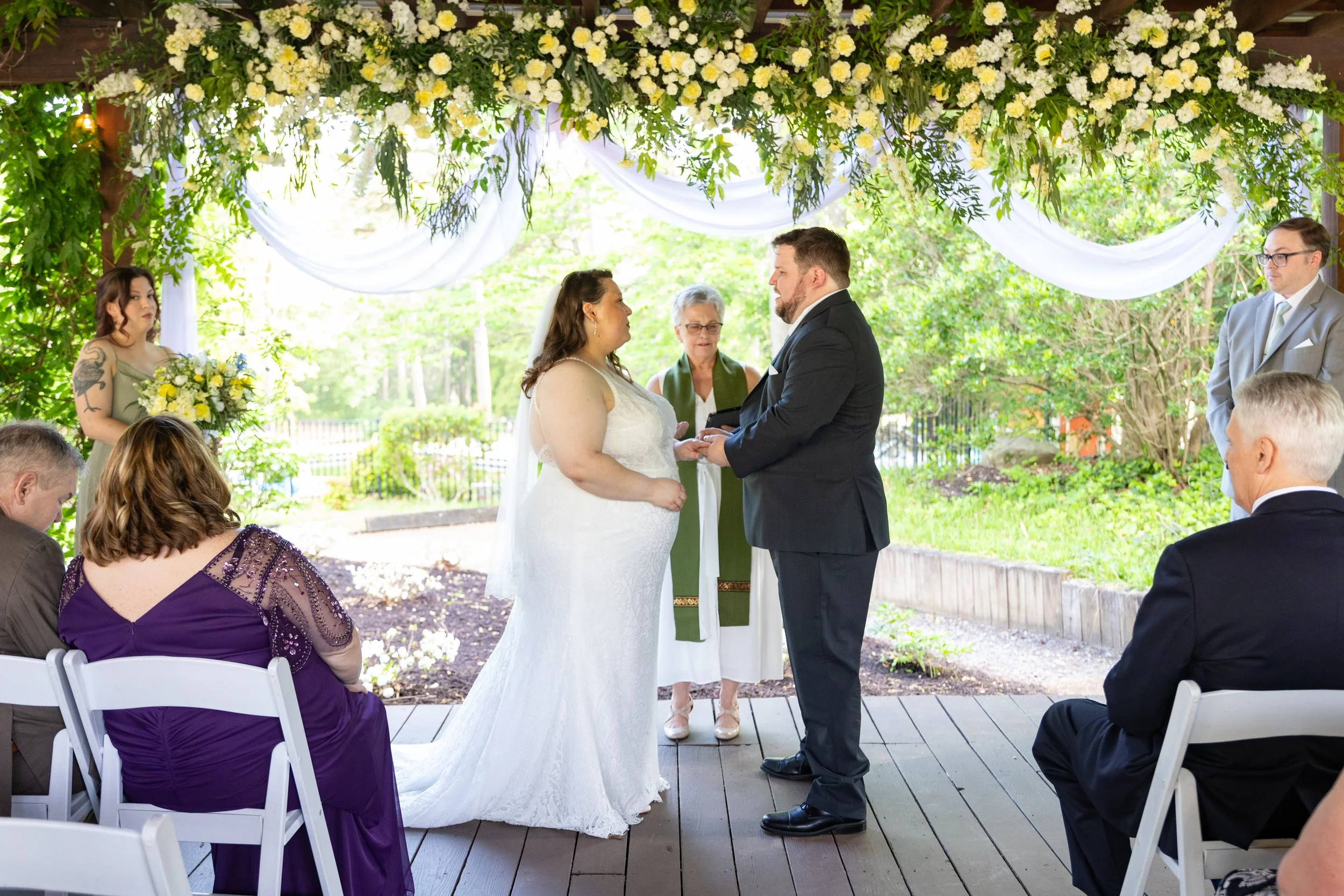 A couple exchanges vows during an outdoor wedding ceremony under a floral canopy with guests seated around them.