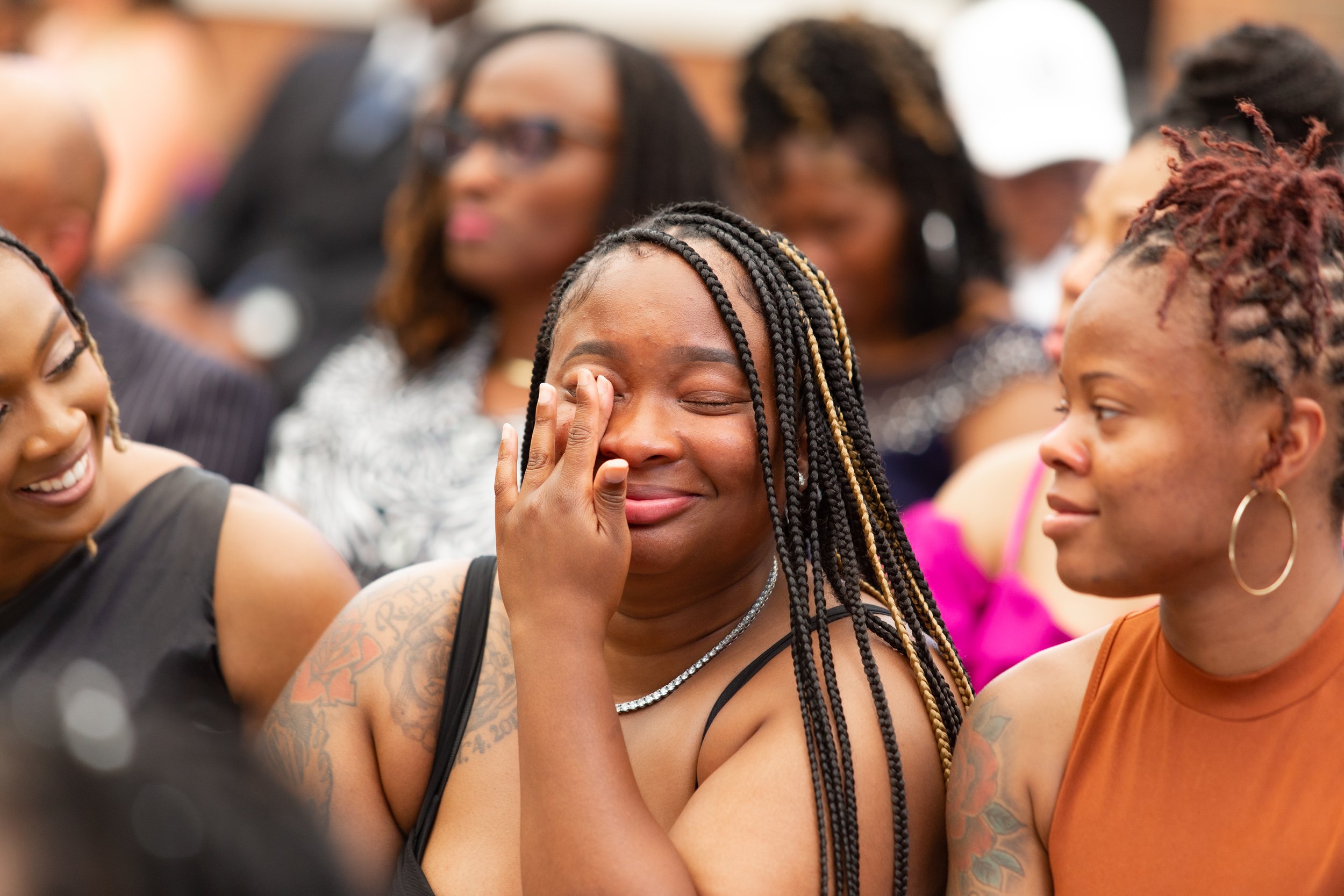Group of diverse women at an outdoor event, with one woman in the center with braided hair laughing and wiping a tear from her eye.