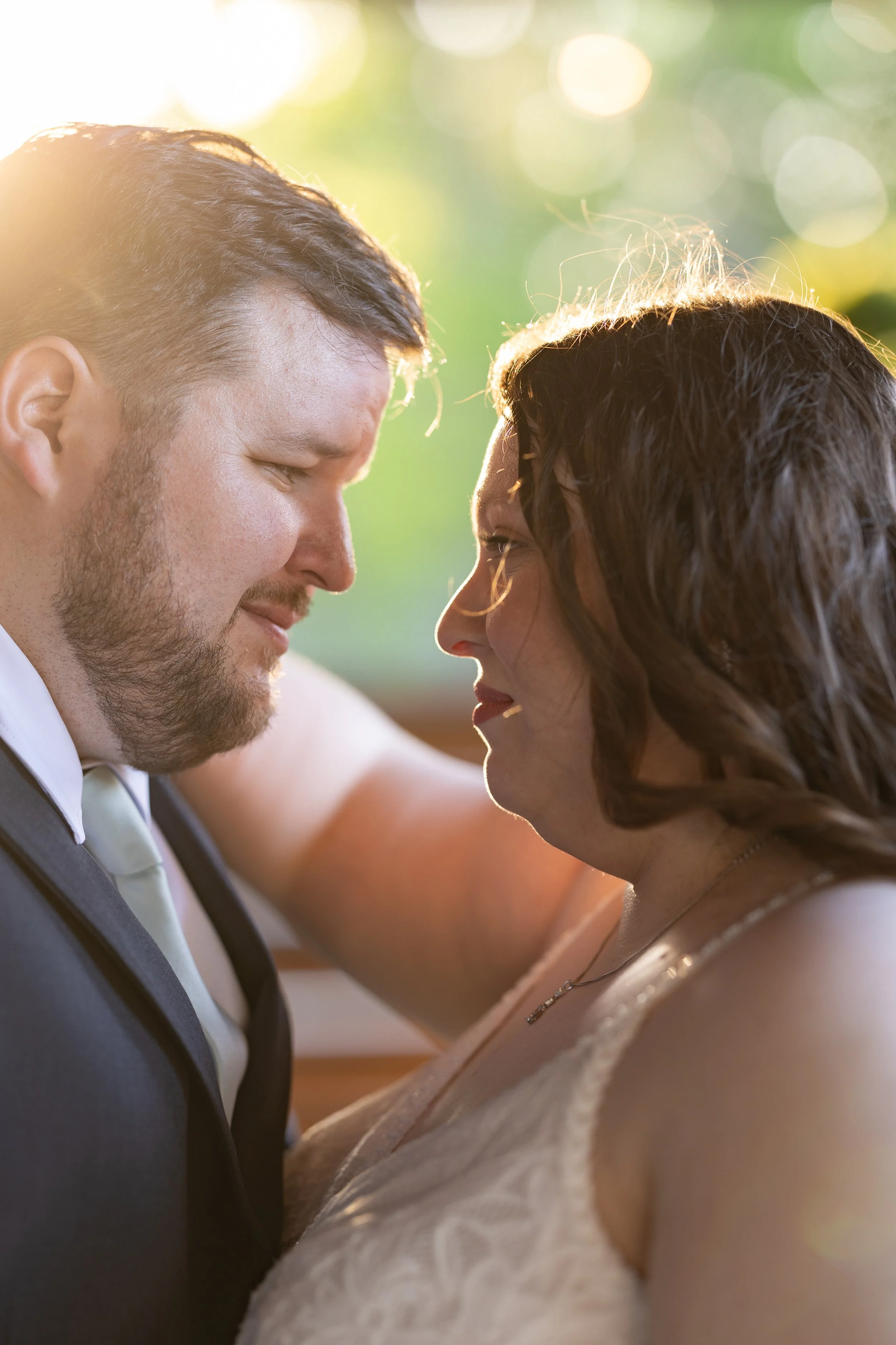 A close-up of a couple touching foreheads, with gentle smiles, outdoors with sunlight and greenery in the background.