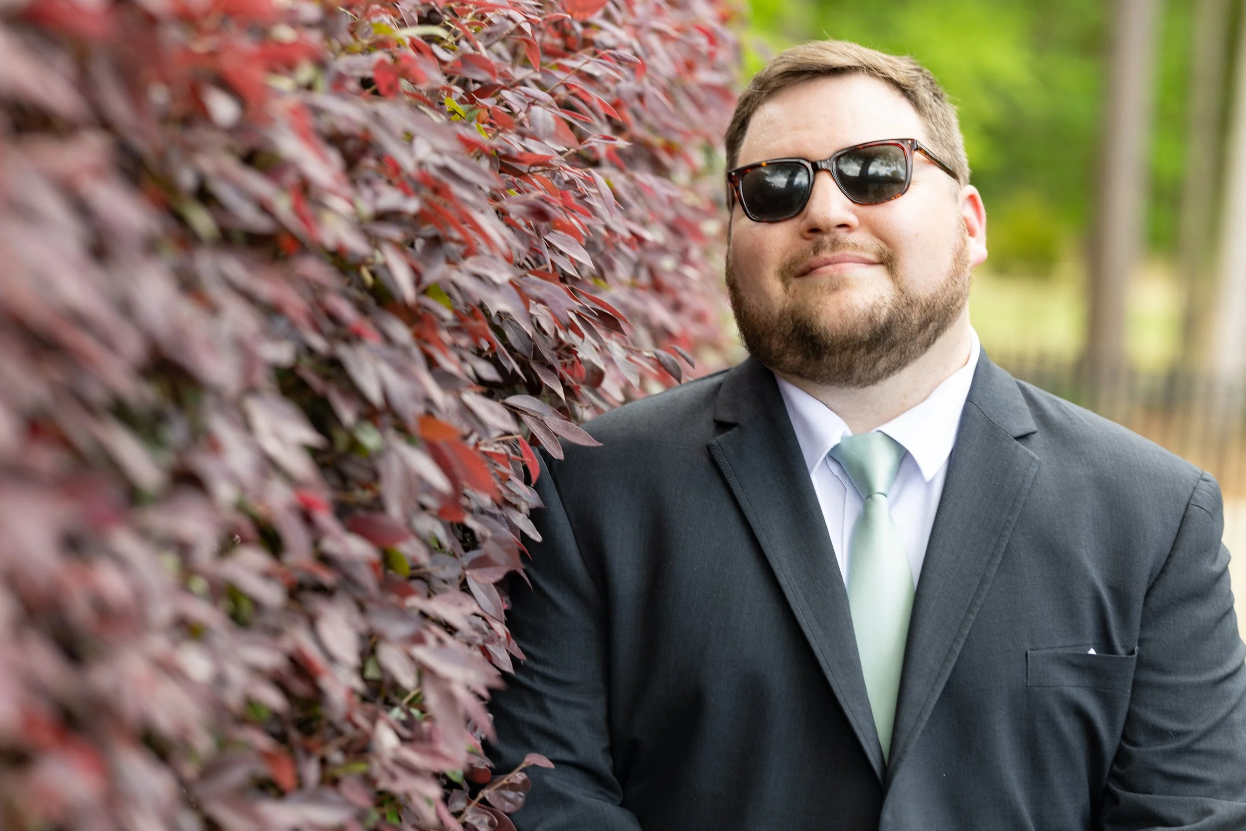 A man in a suit wearing sunglasses standing next to a red and purple leafy hedge, smiling.