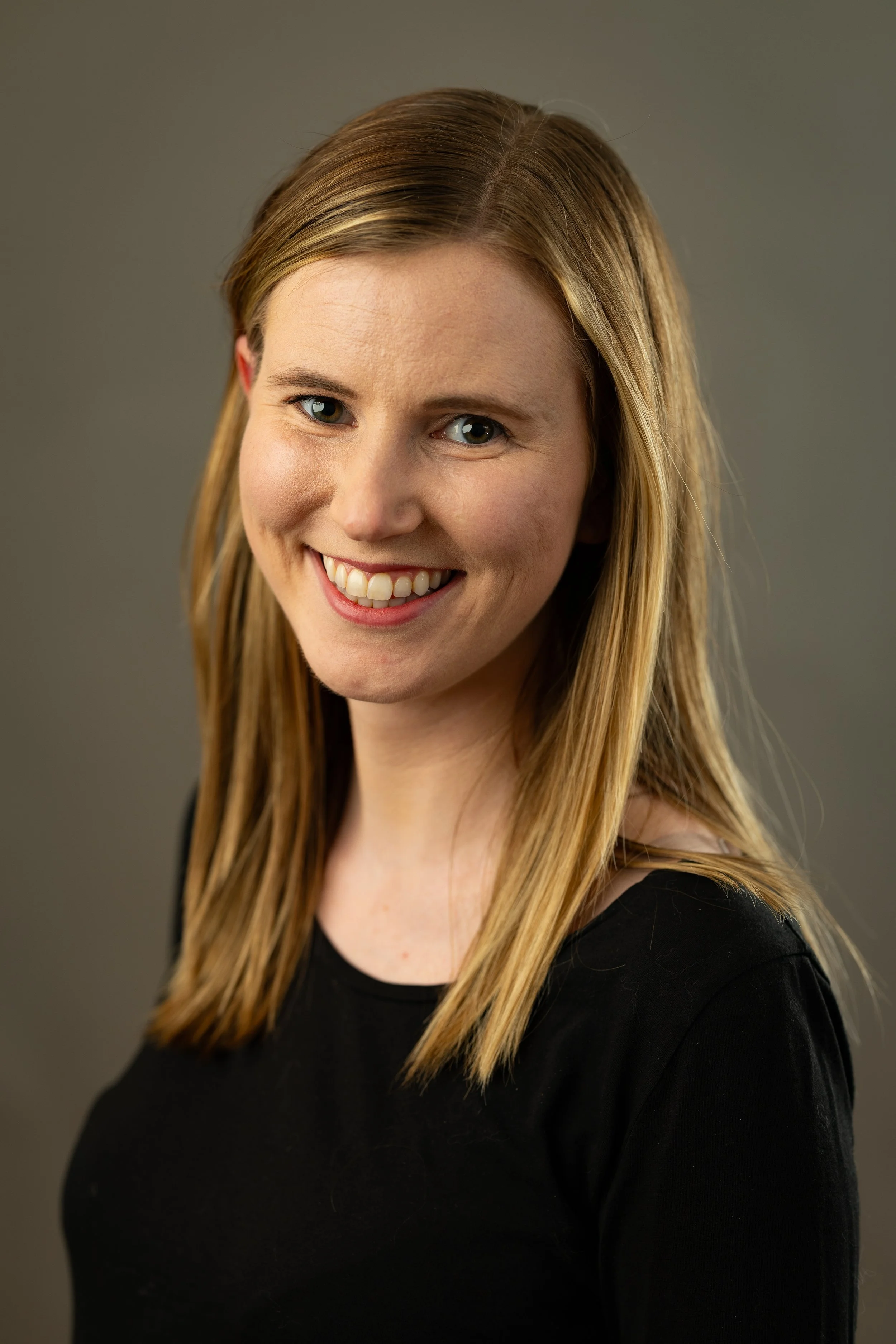 A woman with long blonde hair smiling, wearing a black top, against a neutral background.
