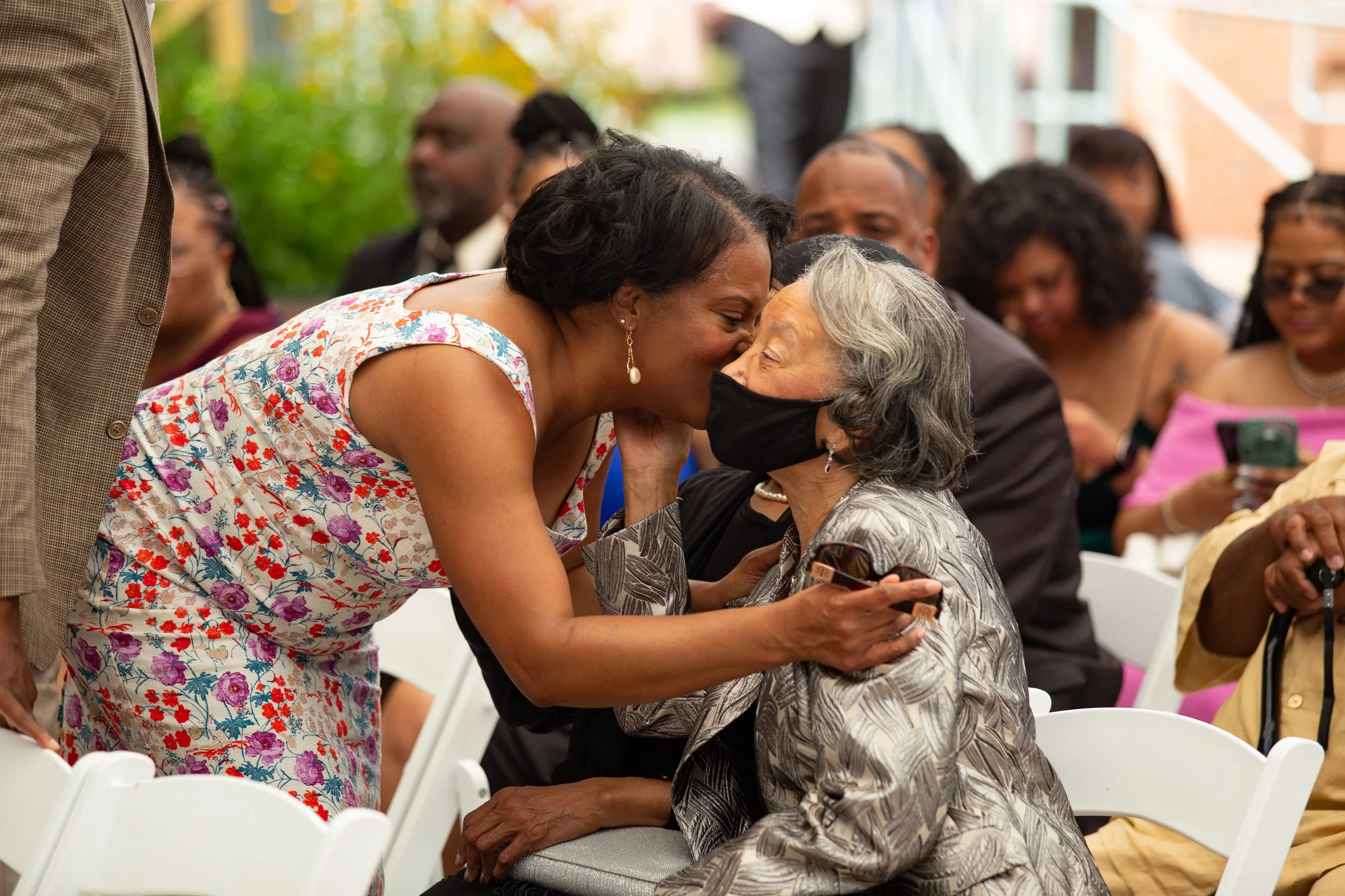 A woman leaning in to kiss an elderly woman on the cheek, both wearing black face masks, at an outdoor gathering with people in the background seated in white chairs.