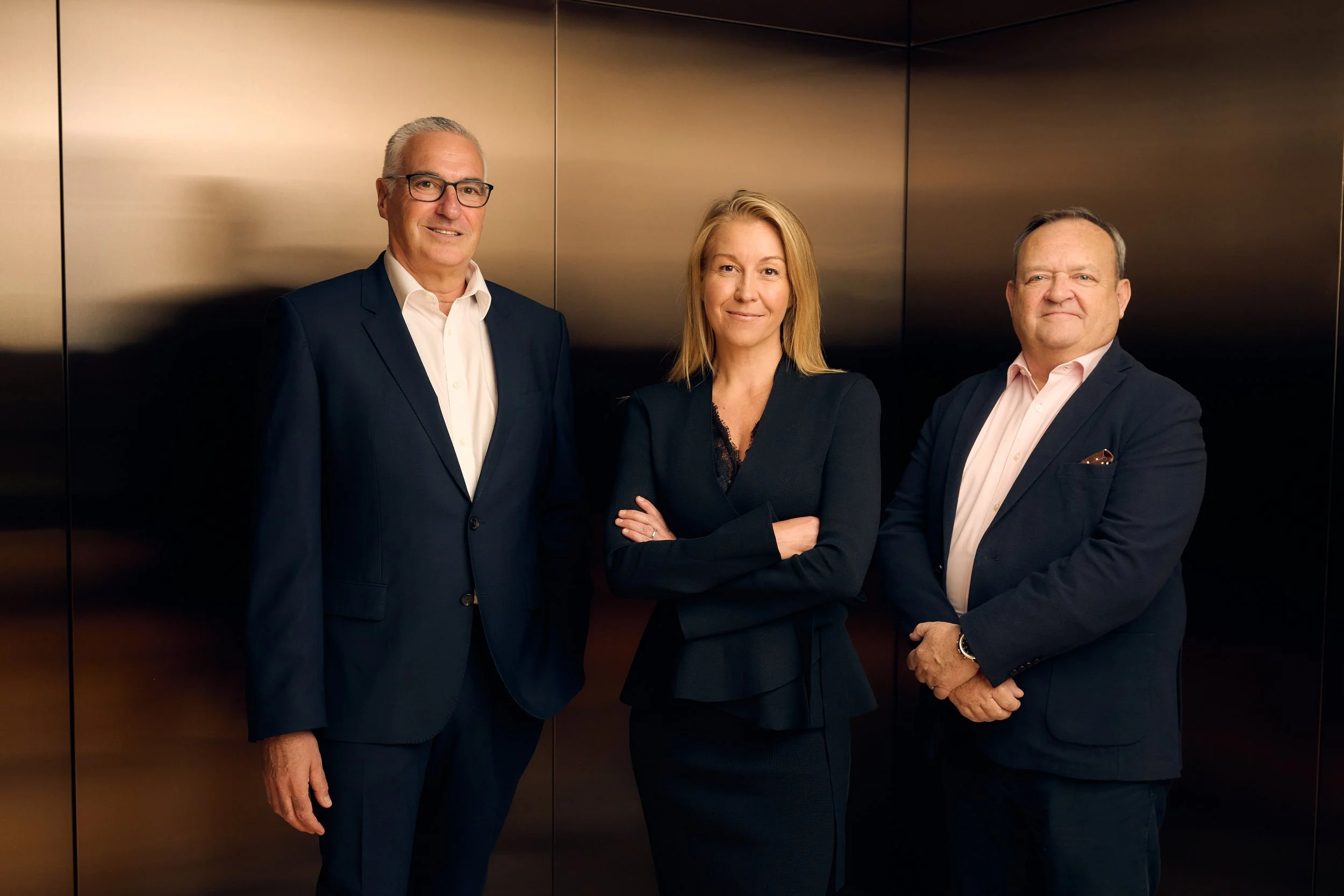 Three professionally dressed individuals standing against a dark, reflective wall, with a man on the left wearing glasses, a woman in the middle with crossing arms, and a man on the right with hands clasped.