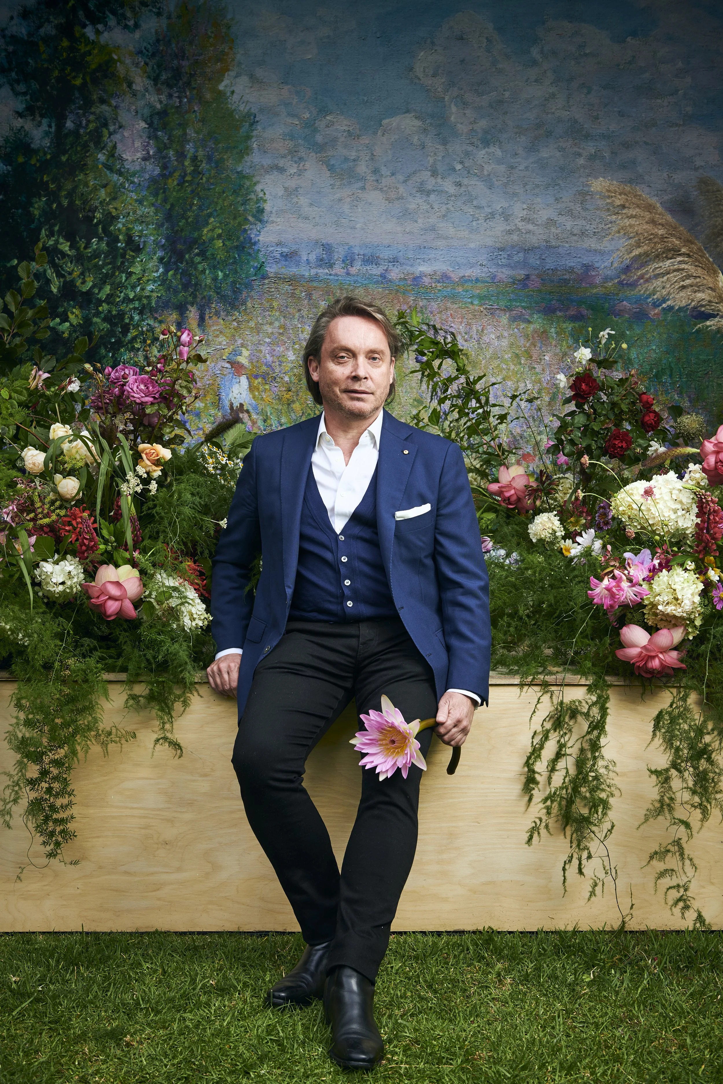 A man in a blue suit sitting in front of a backdrop of a landscape painting and a lush flower arrangement, holding a pink water lily blossom and a pair of sunglasses.