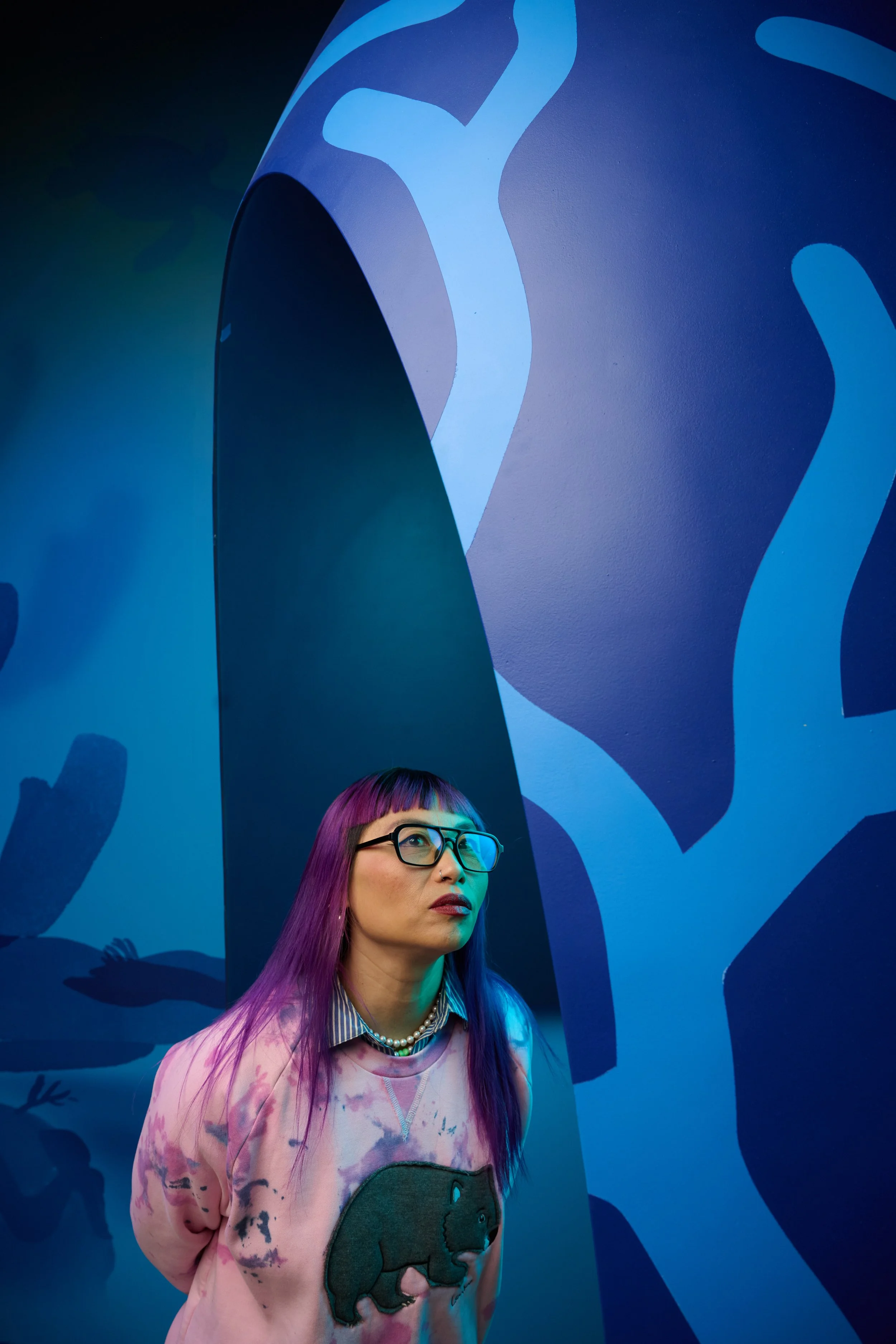 A woman with purple hair and glasses standing against a blue abstract patterned wall.