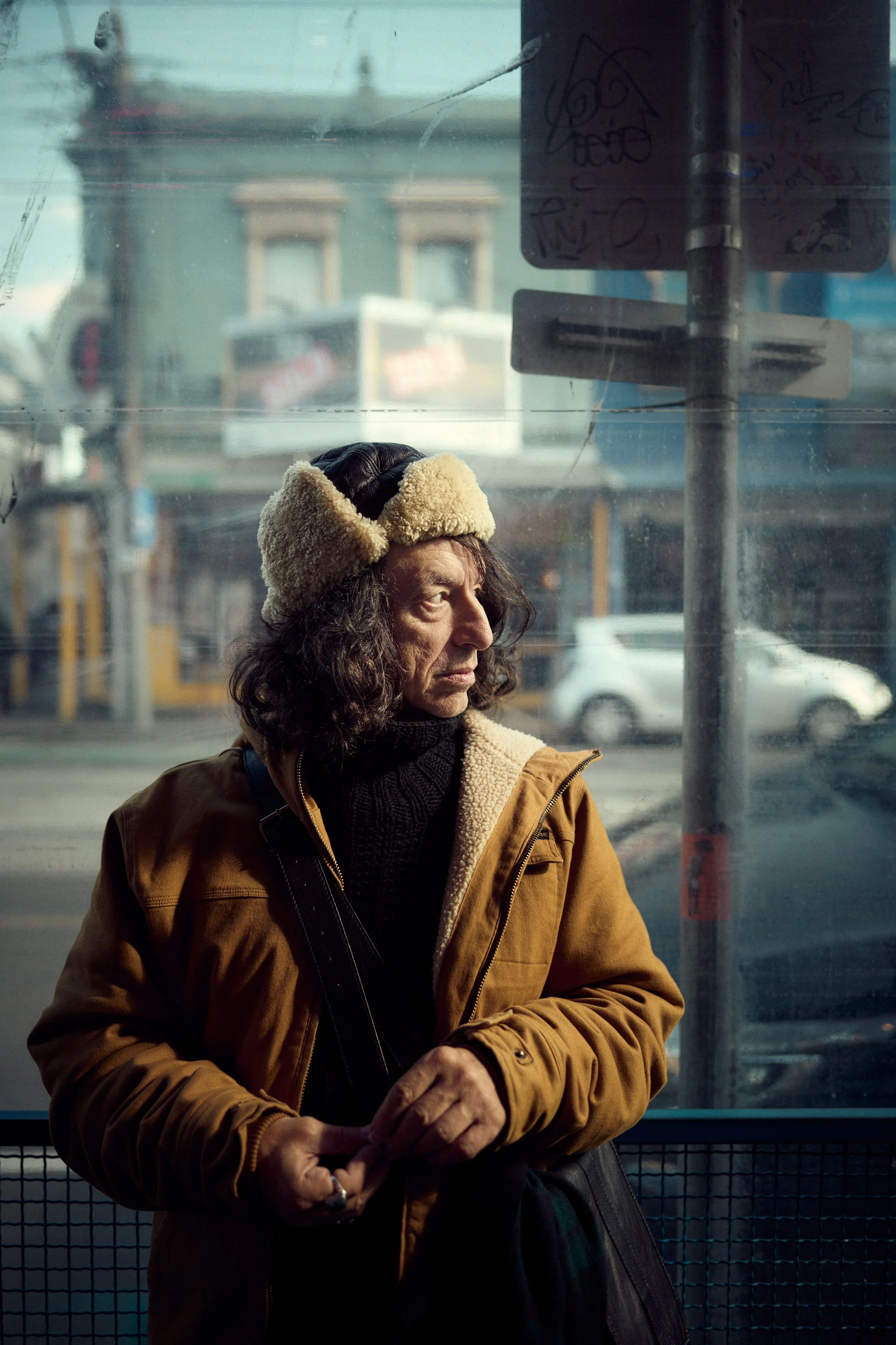 A woman with long curly hair wearing a brown jacket and a beige furry hat with ear flaps, standing at a bus stop or on a sidewalk in an urban area. The background shows a building, a graffiti-covered sign, and some vehicles, indicating a city street 