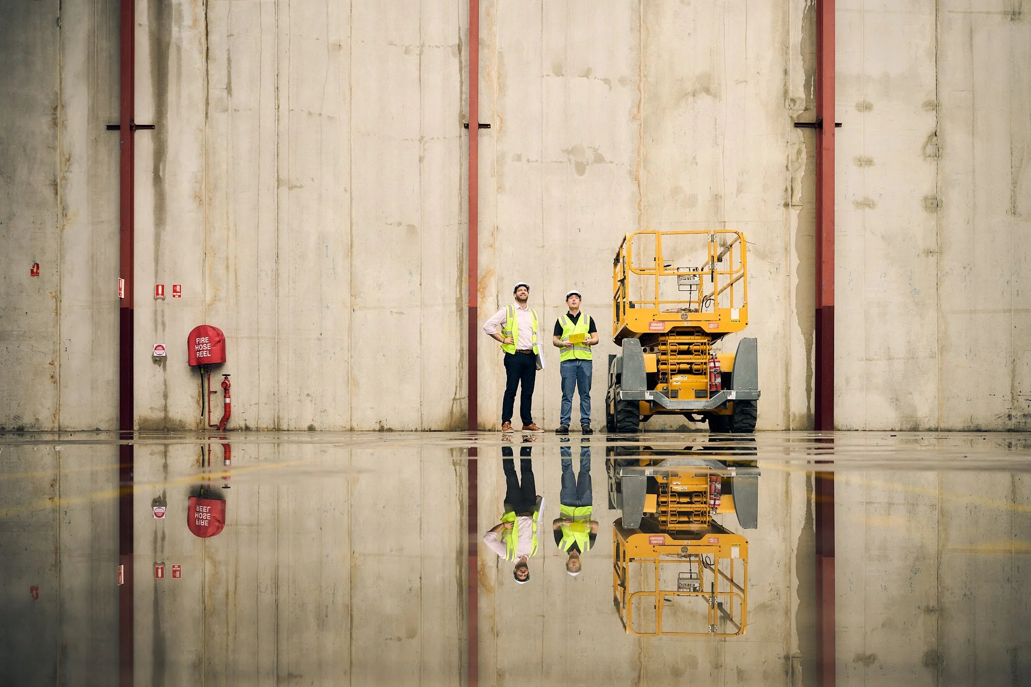 Two workers in safety vests and helmets standing beside a yellow lift inside a large industrial space with a high concrete wall. Their reflection is visible on a shiny floor, with fire safety equipment and pipes attached to the wall.