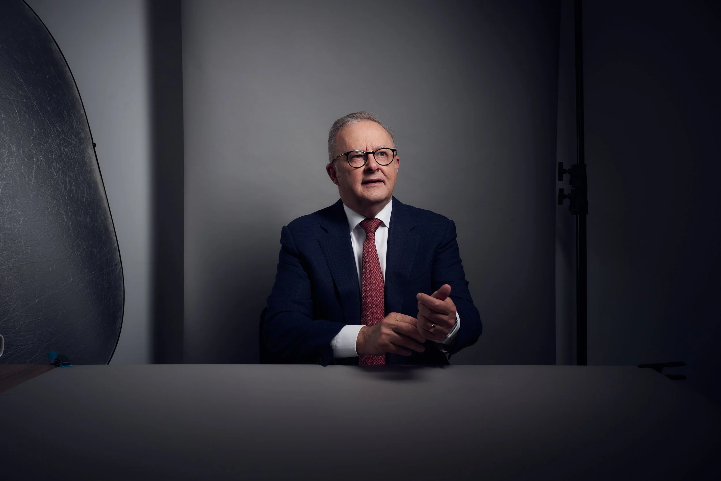 A man in a dark suit and red tie sits at a table with a plain gray background, wearing glasses and looking slightly to his left with a thoughtful expression.