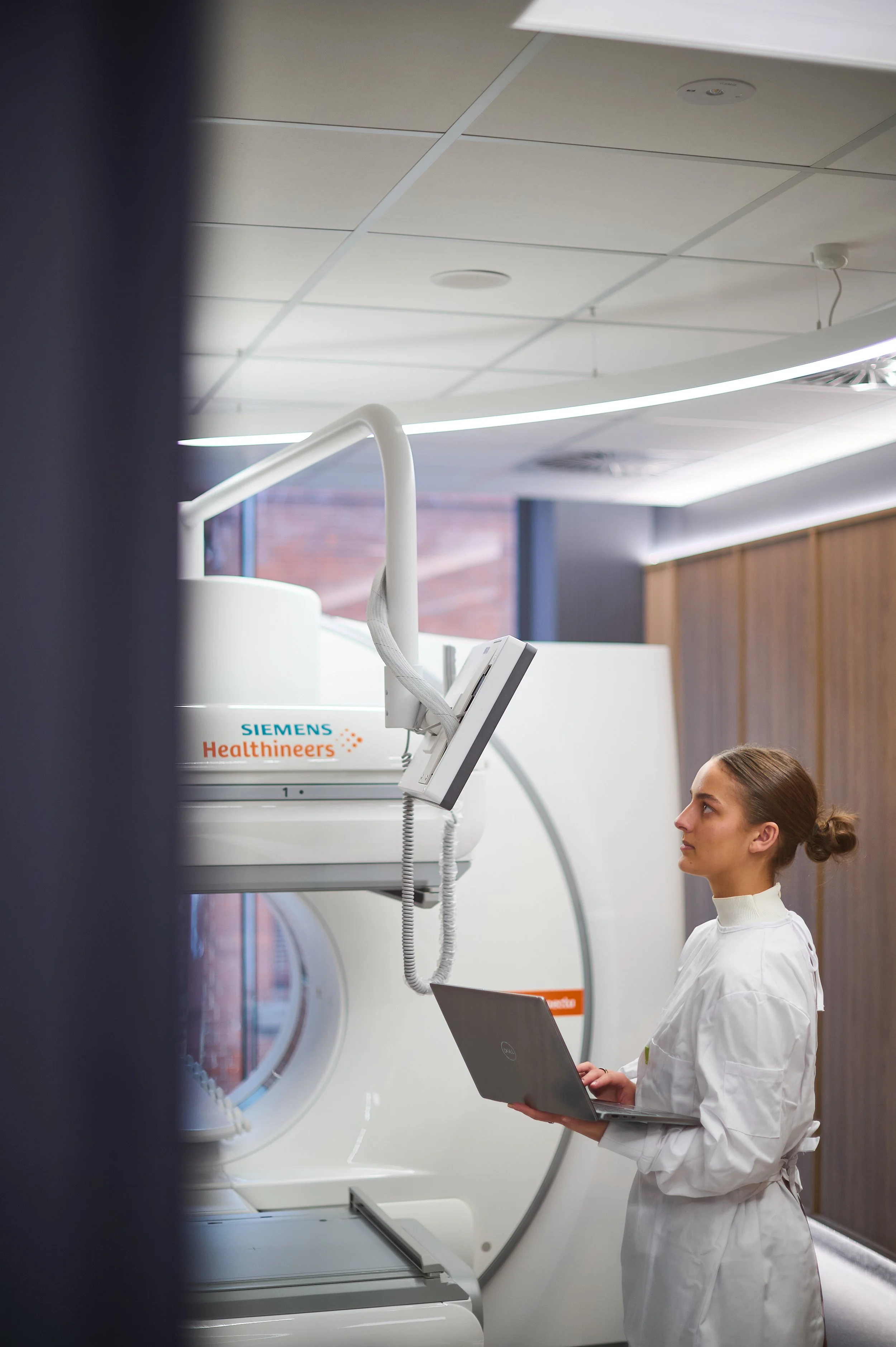 A female medical professional in a white lab coat standing next to a Siemens Healthineers MRI machine, holding a laptop, inside a modern clinical setting.