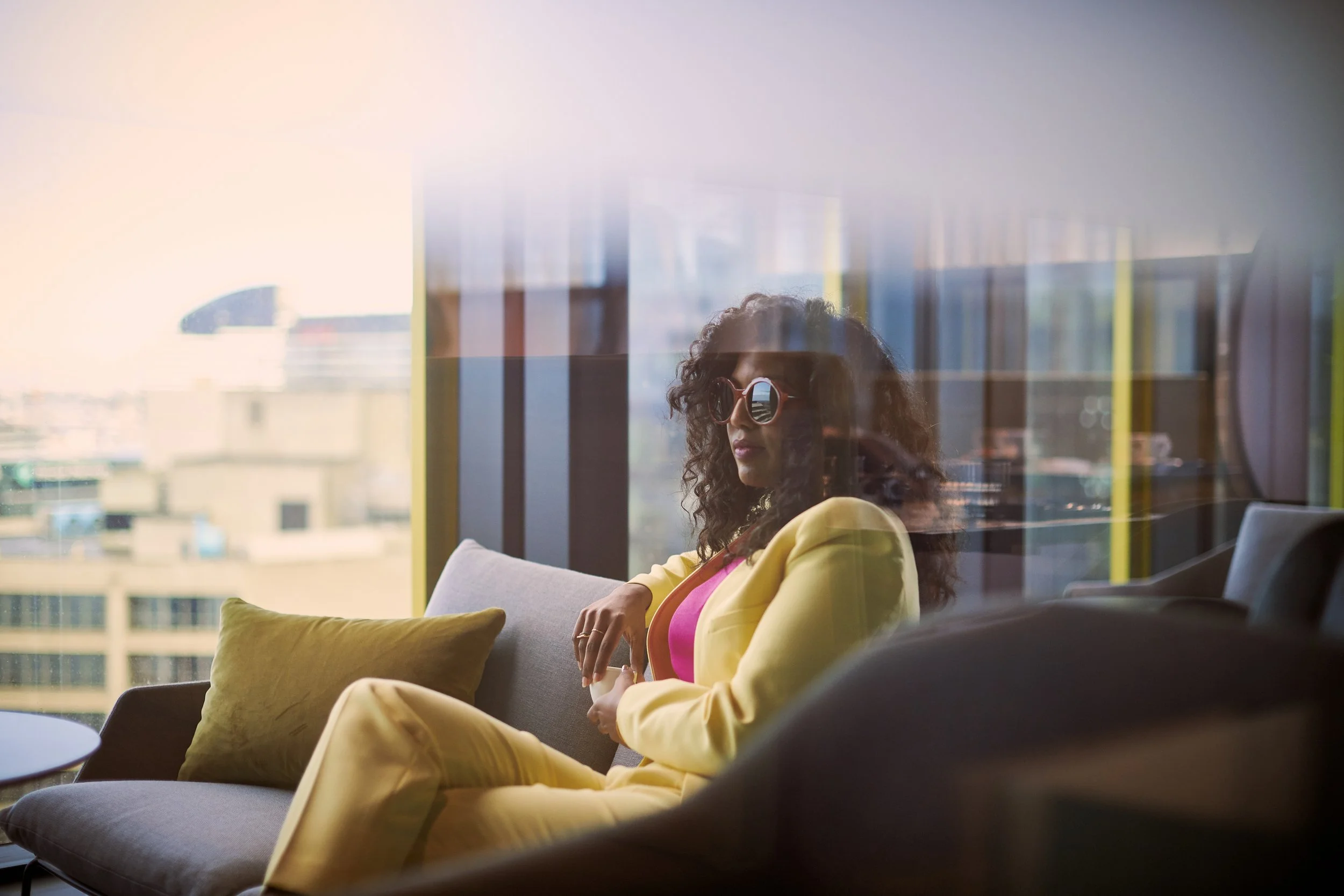 A woman with curly hair wearing sunglasses and a yellow blazer sitting on a sofa in a bright room with large windows, looking outside.