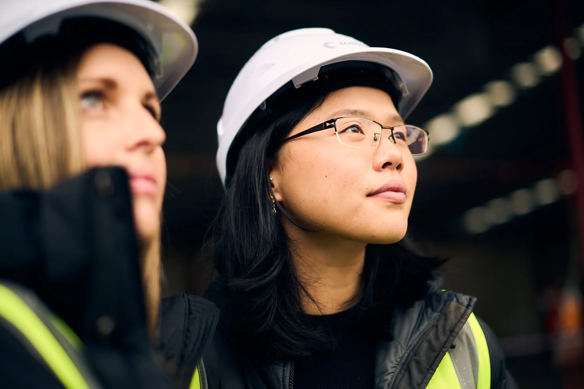 Close-up of two construction workers wearing white hard hats and high-visibility yellow vests, standing outdoors.